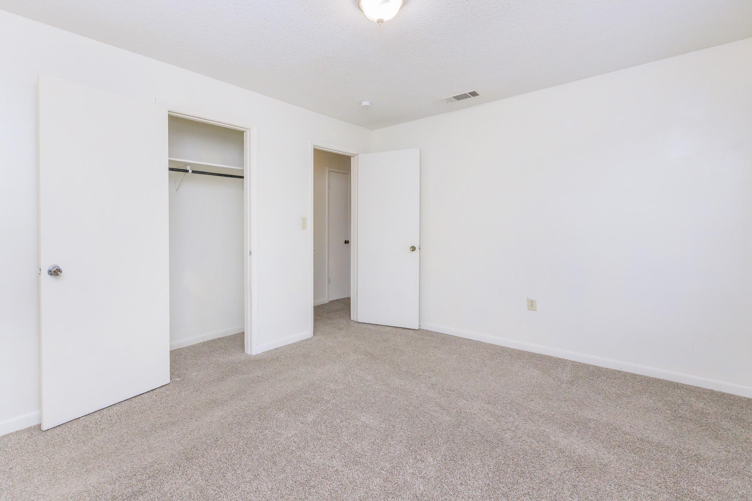 Empty room with light-colored walls and beige carpeting. Two open white doors lead to closets, and a ceiling light fixture is visible. The space is well-lit and has a clean, minimalistic appearance, suitable for furnishing as a bedroom or office.