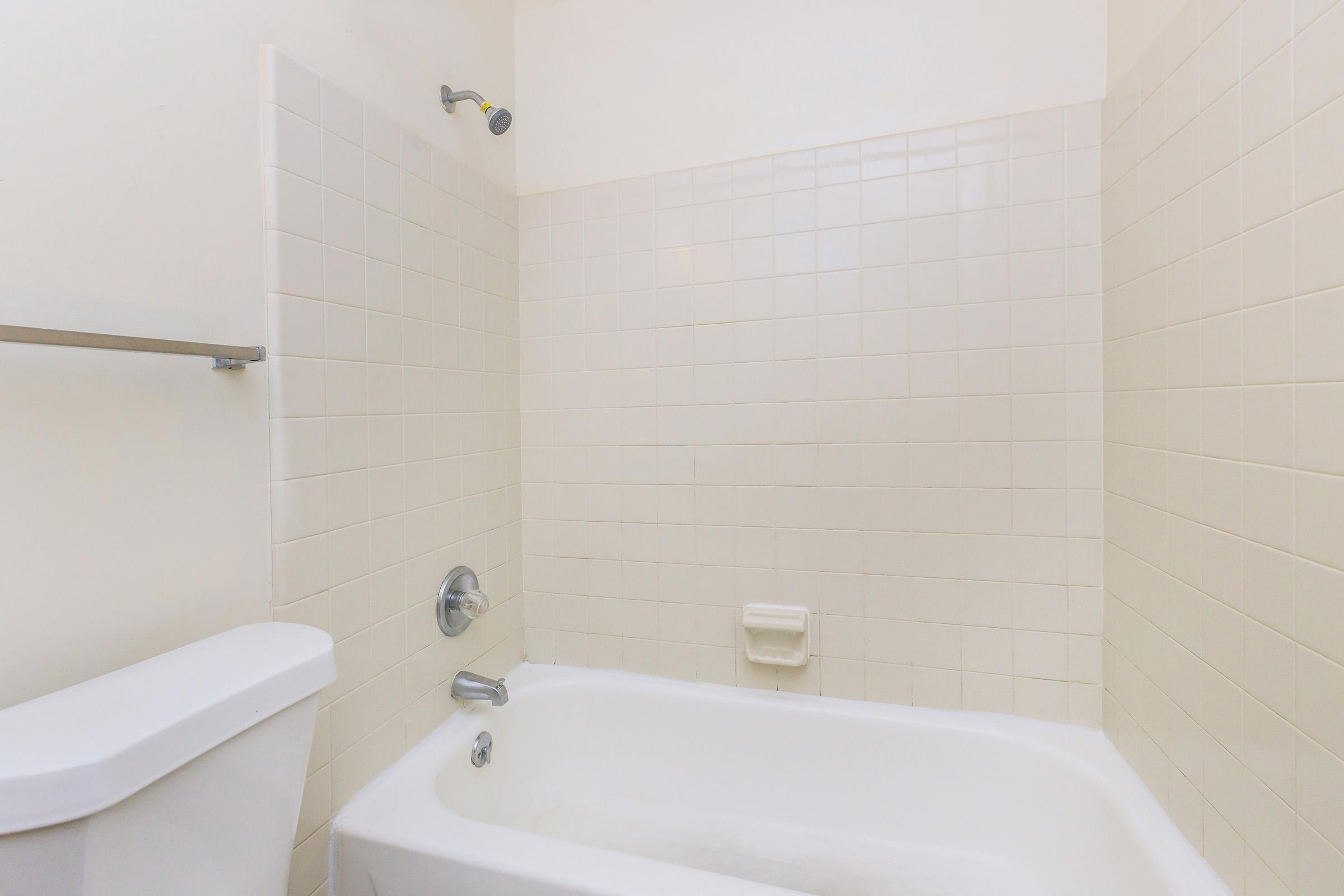 A clean, minimalistic bathroom featuring a white bathtub, showerhead, and a white toilet. The walls are tiled in light-colored squares, and there is a towel bar mounted on the wall. The overall look is bright and simple, suitable for a functional bathing space.