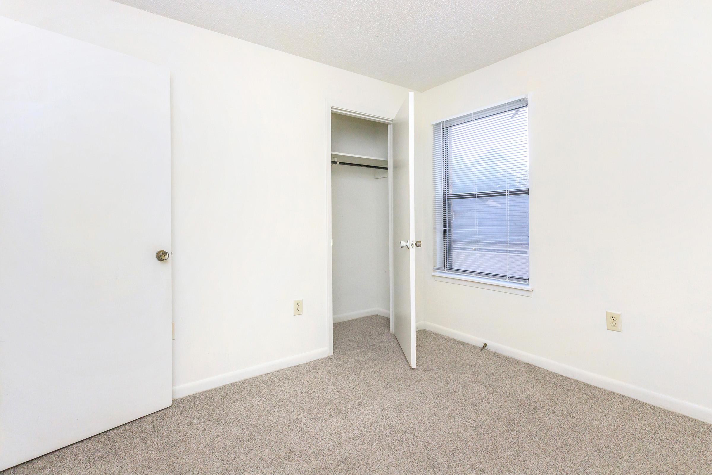 A bright, empty room featuring light-colored walls, a carpeted floor, a closet with an open door on the left, and a window with blinds on the right, providing natural light.