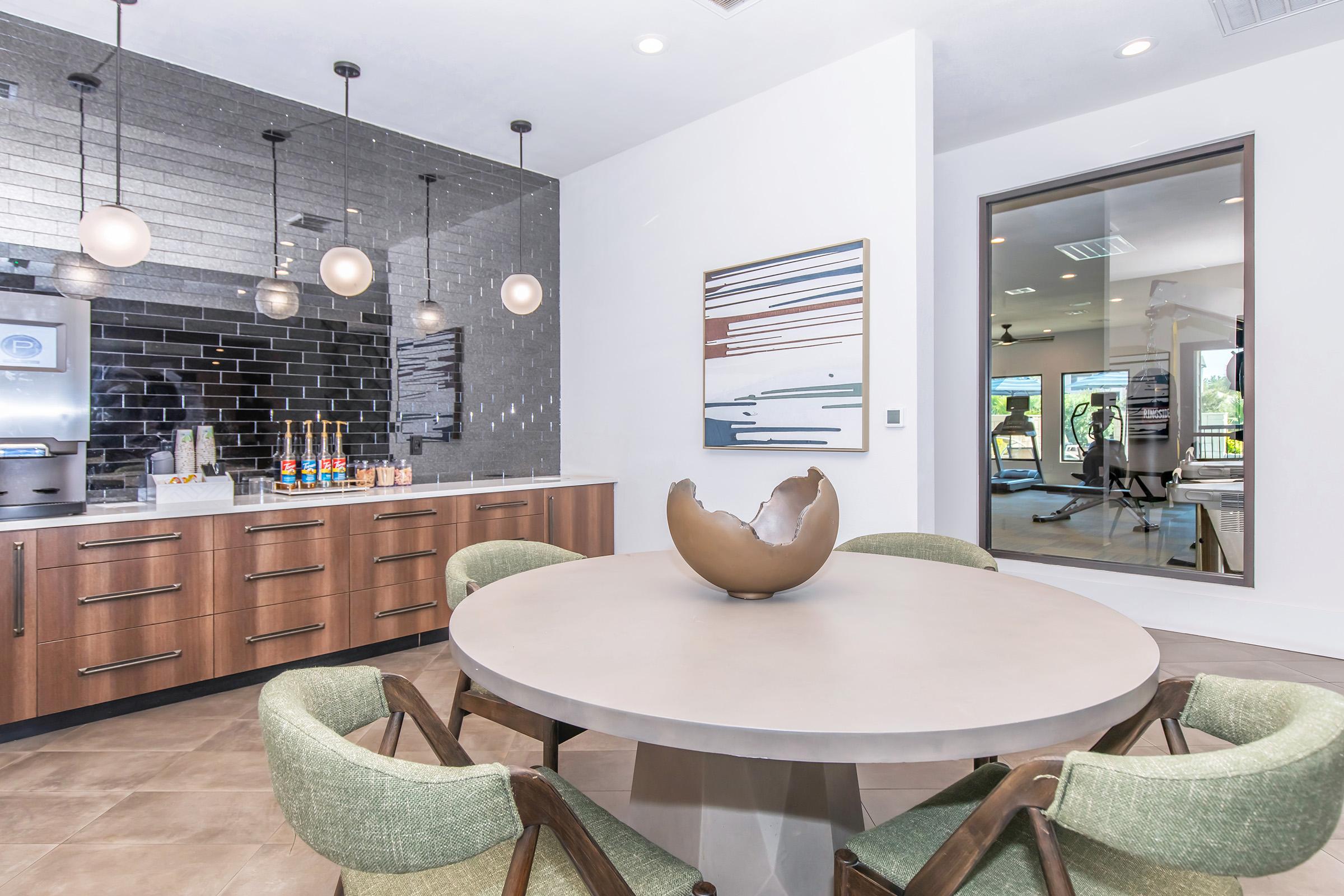 A modern kitchen and dining area featuring a round table with a large decorative bowl at its center. The cabinetry is wooden with a sleek finish, complemented by a stylish backsplash of rectangular tiles. Soft lighting hangs above, and a mirror reflects a fitness area in the background.