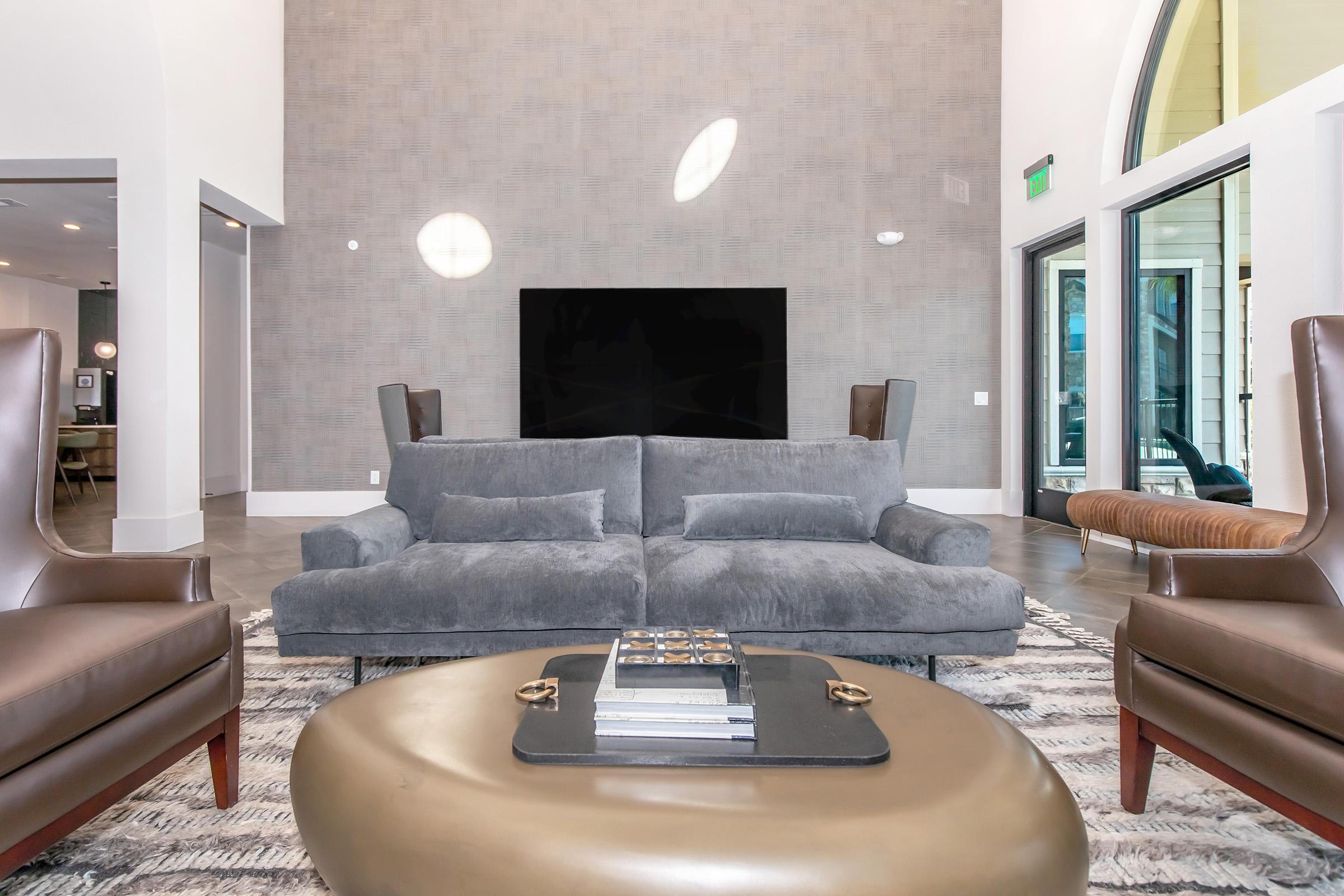A modern living room featuring a grey sofa centered in front of a large flat-screen TV mounted on a textured wall. Two dark brown armchairs flank the sofa, and a round, gold coffee table sits on a patterned rug. Large windows allow natural light to enter the space, enhancing the contemporary decor.