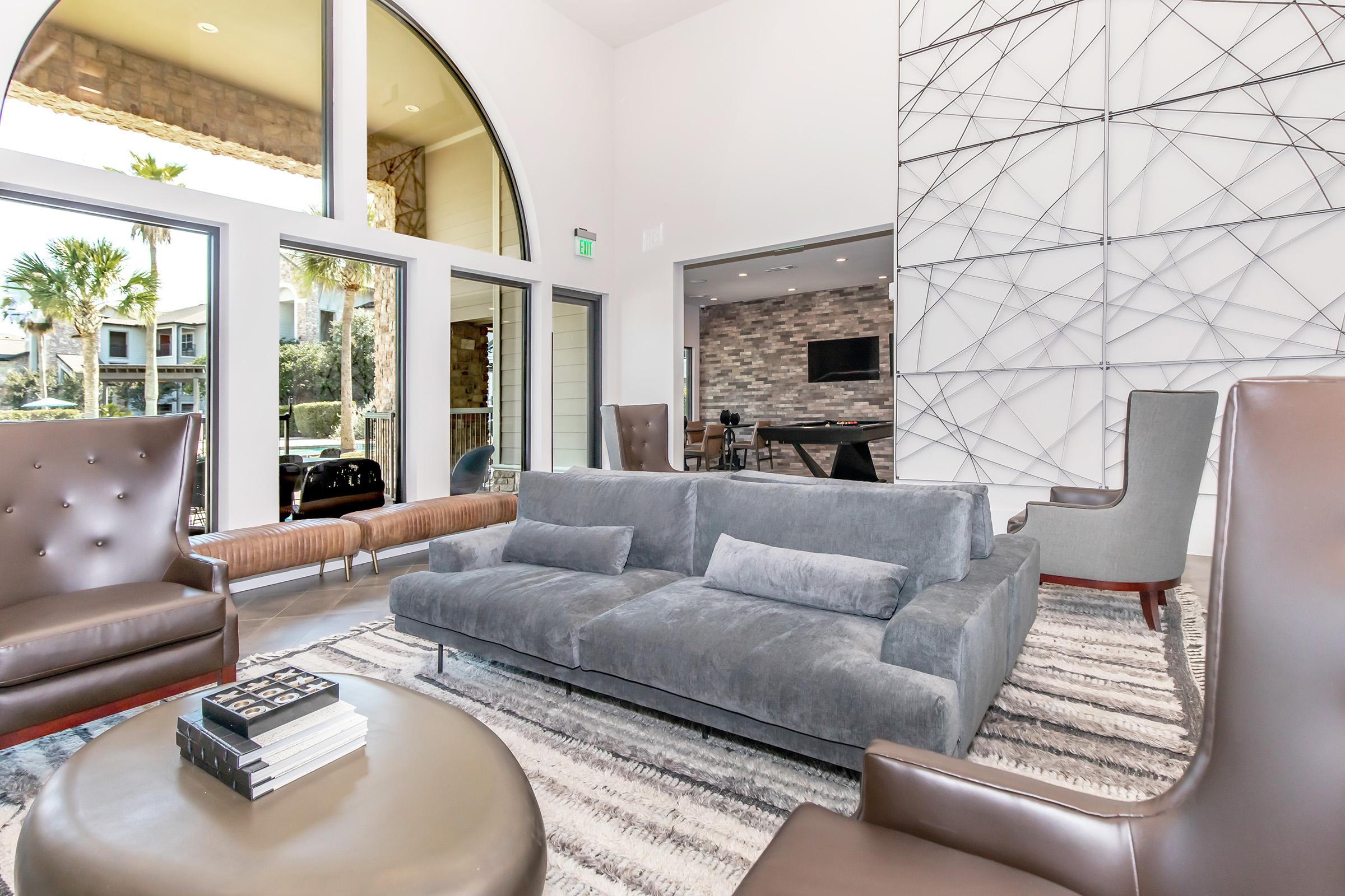 A modern lounge area featuring a gray sectional sofa with cushions, two brown leather armchairs, and a round coffee table with books on it. Large windows allow natural light, and a geometric wall art adds a stylish touch. A hint of greenery is visible outside, enhancing the inviting atmosphere.