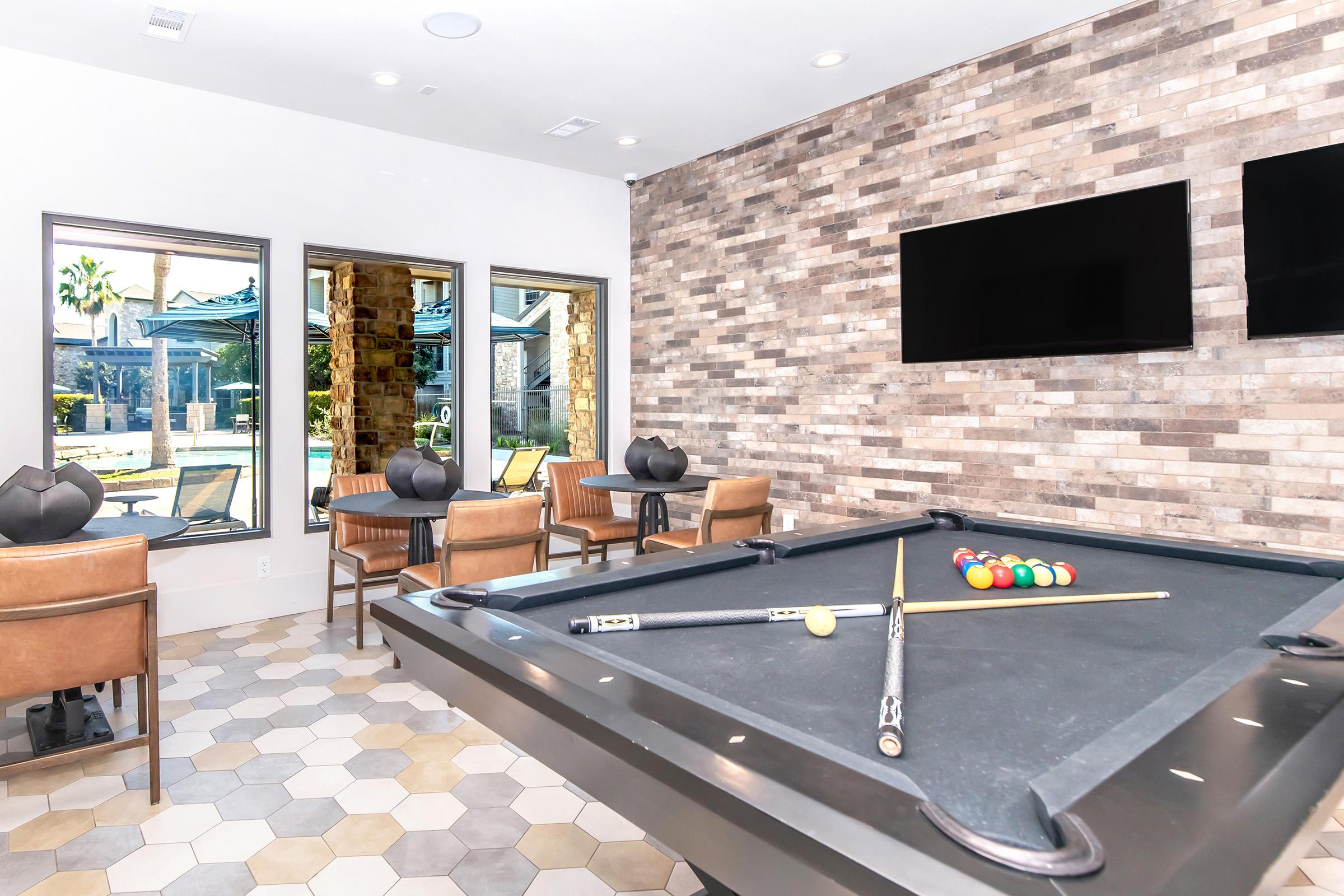 A stylish lounge area featuring a billiard table with pool cues and colored balls, surrounded by comfortable seating. Large windows provide a view of an outdoor pool area, and a modern brick wall adds texture to the elegant space. Two flat-screen TVs are mounted on the wall.