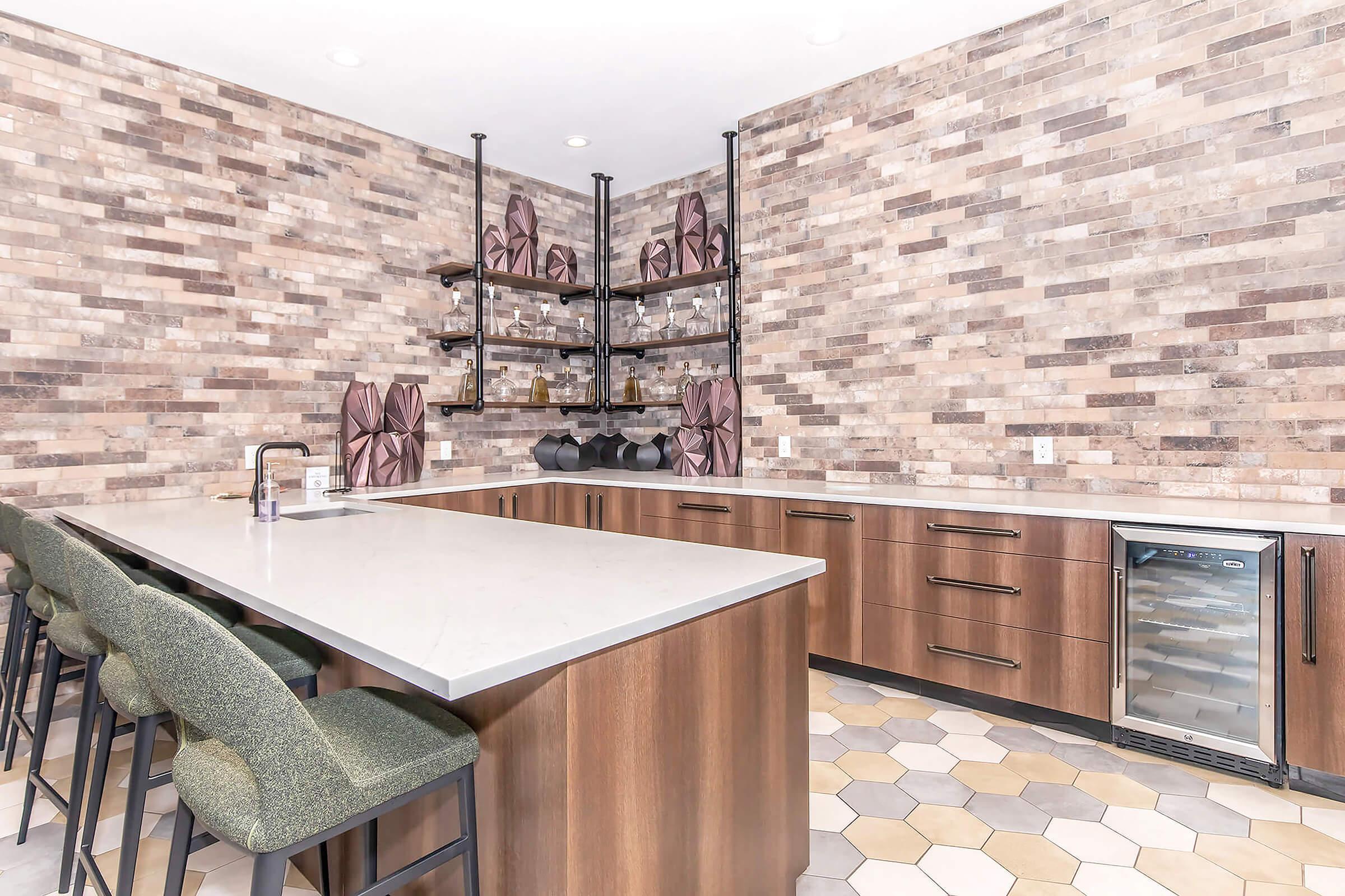 Modern kitchen featuring a large island with a white countertop, dark wooden cabinetry, and a decorative wall of textured bricks. Shelves hold stylish glassware and decorative pieces. Bar stools in green fabric line the island, and there is a built-in wine cooler. The floor is adorned with hexagon tiles in neutral tones.