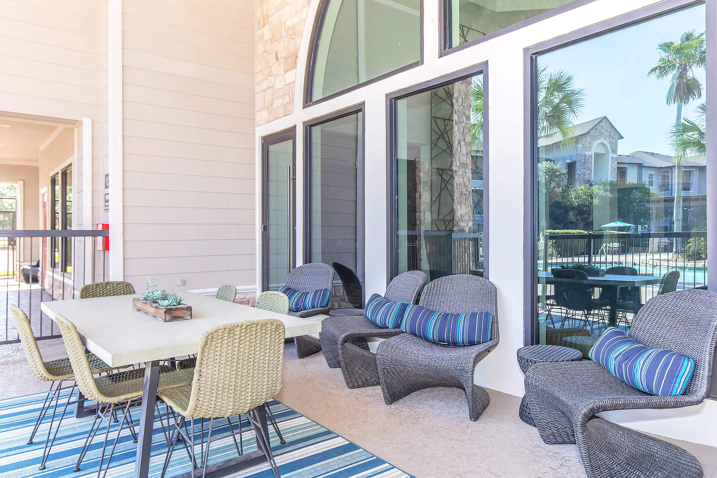 A modern outdoor lounge area featuring comfortable wicker furniture with cushions, a table, and a decorative rug. Large windows provide views of a pool area surrounded by palm trees and greenery, creating a relaxing and inviting atmosphere.