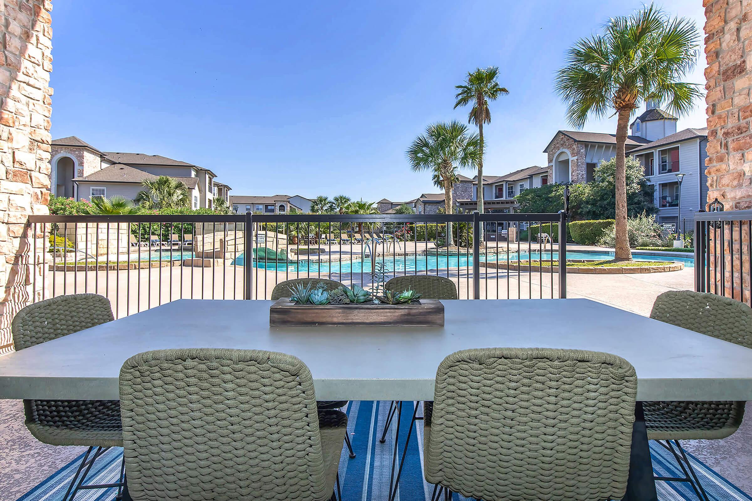 A spacious outdoor dining area featuring a modern table with green chairs, positioned near a pool surrounded by palm trees and apartment buildings. The bright blue sky enhances the inviting atmosphere of the space, suggesting a relaxing outdoor environment.