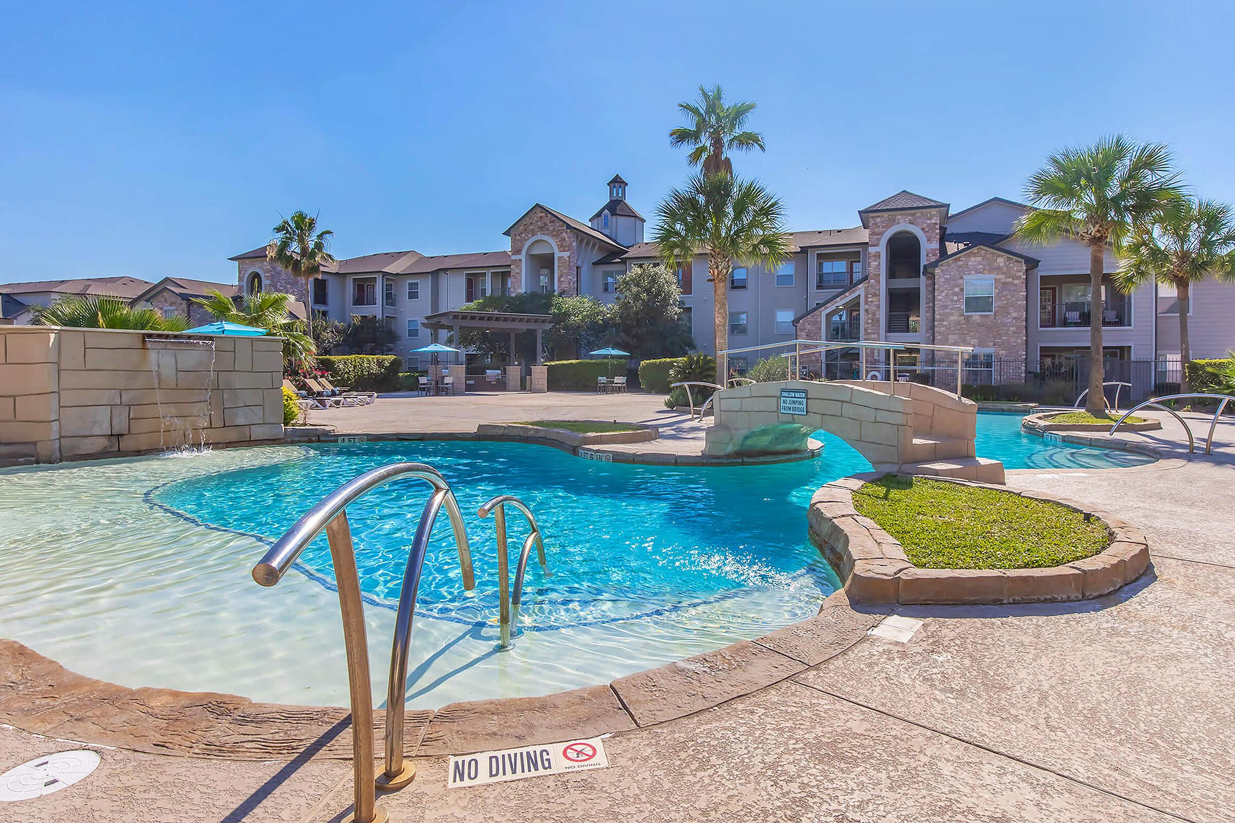 A resort-style swimming pool with a gentle curve, surrounded by lush landscaping and palm trees. The pool features a decorative bridge and a "No Diving" sign. In the background, an apartment complex is visible under a clear blue sky.