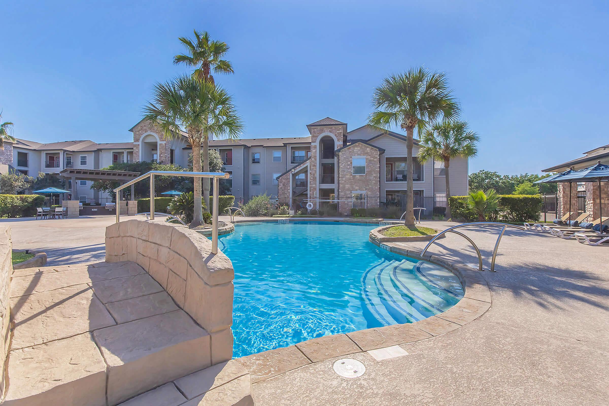 A sunny outdoor pool area surrounded by palm trees and lounge chairs, with a modern apartment building in the background. The pool features a curved shape and a stone pathway leading to it, creating a relaxed and inviting atmosphere. Clear blue sky enhances the tranquil scene.