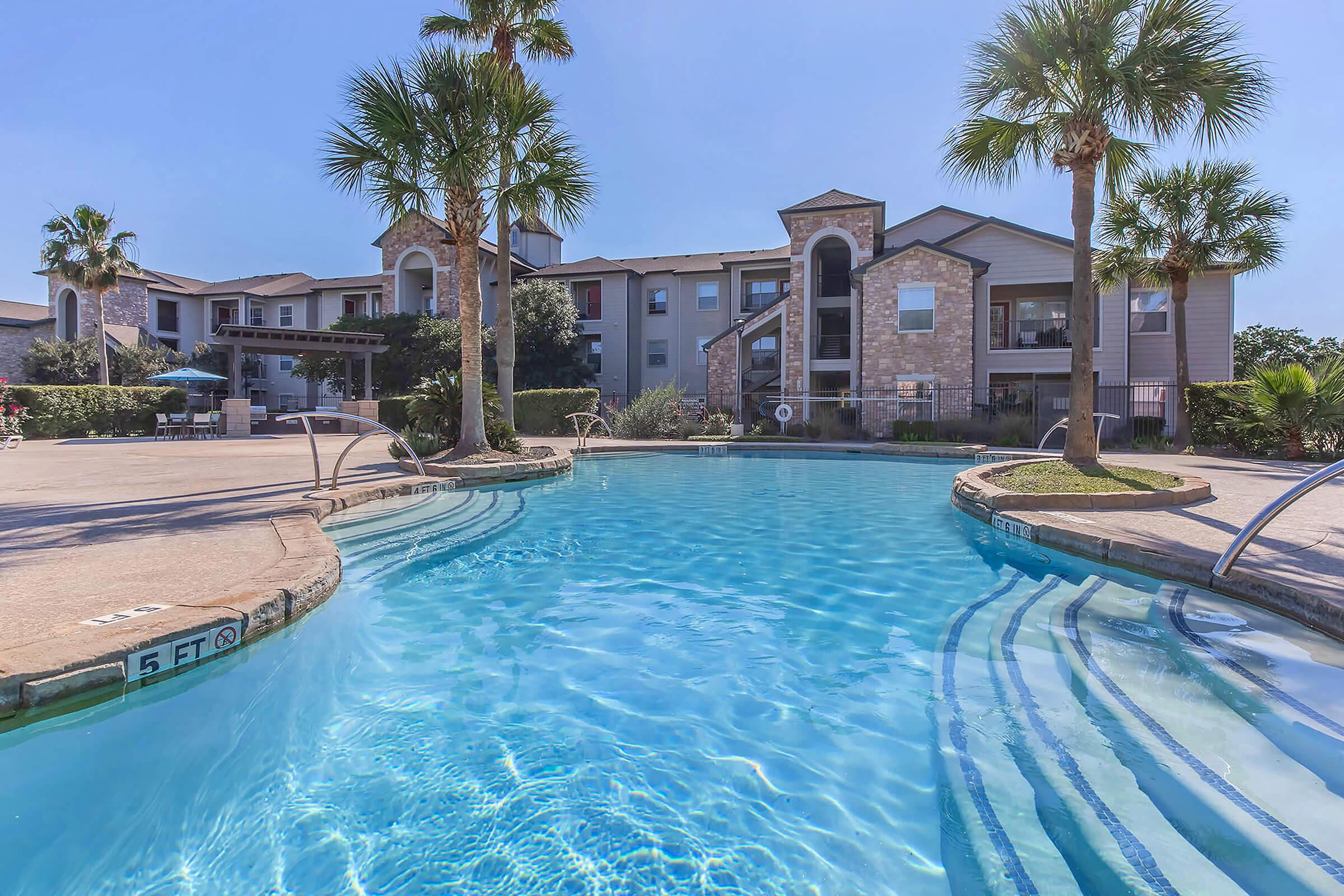 A sparkling swimming pool with clear blue water surrounded by palm trees. In the background, there are residential buildings with stone and wood exteriors, along with a poolside area featuring lounge chairs and umbrellas. The setting appears inviting and well-maintained, under a bright blue sky.
