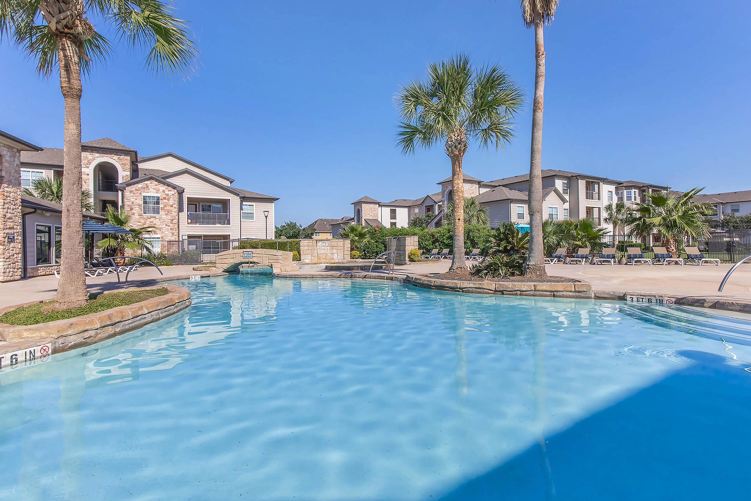 A sunny outdoor pool area surrounded by palm trees and lounge chairs. In the background, there are modern apartment buildings with balconies. The water is clear and inviting under a blue sky, creating a relaxing atmosphere.