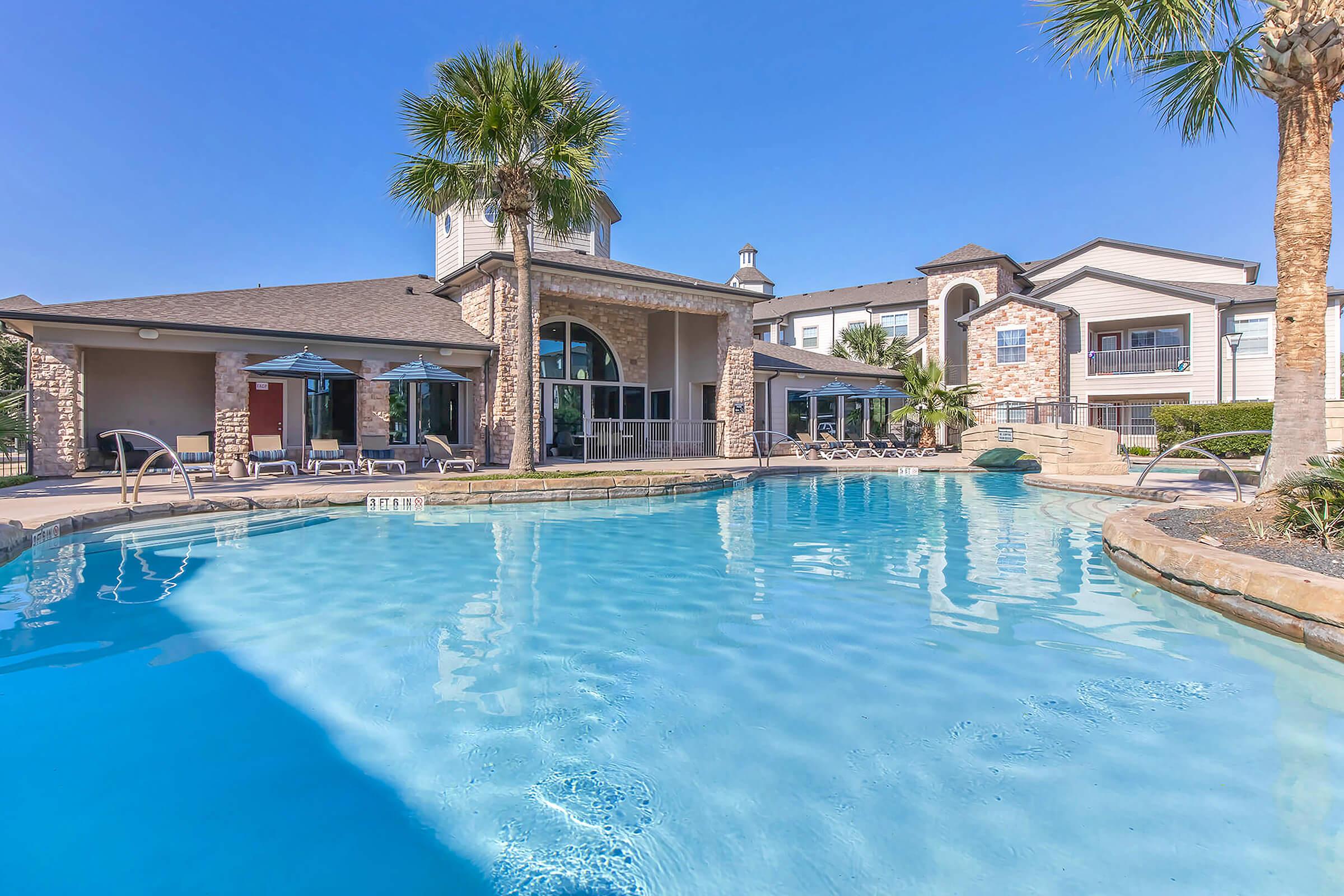 A vibrant outdoor swimming pool with clear blue water, surrounded by palm trees and lounge chairs. In the background, a modern building features large windows and a stylish design, under a clear blue sky. Perfect for relaxation and leisure in a sunny environment.