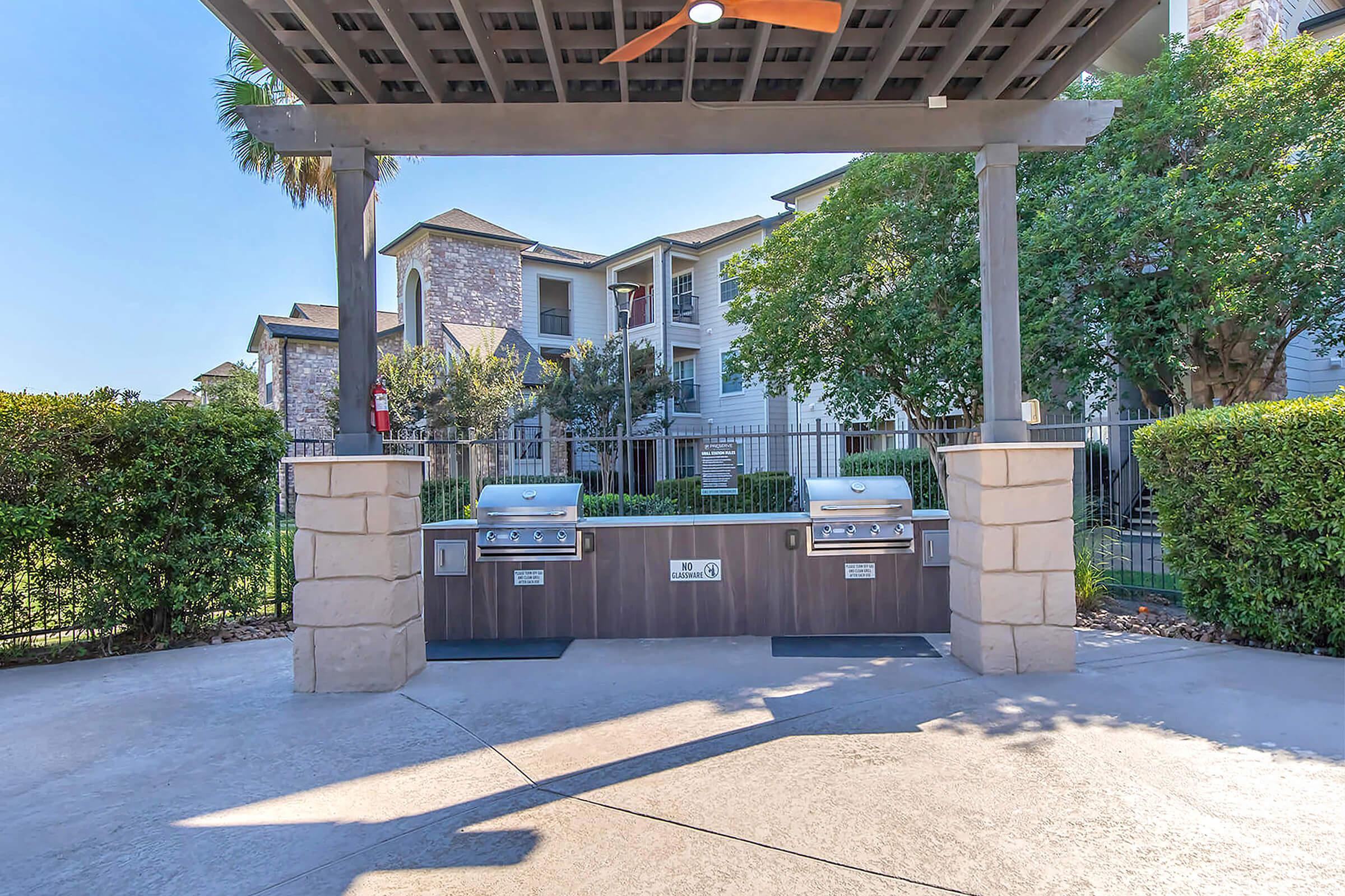 Outdoor grilling area under a shaded pergola, featuring two stainless steel grills, surrounded by lush greenery and apartment buildings in the background. The scene captures a sunny day with a clear blue sky, creating a welcoming space for outdoor cooking and social gatherings.