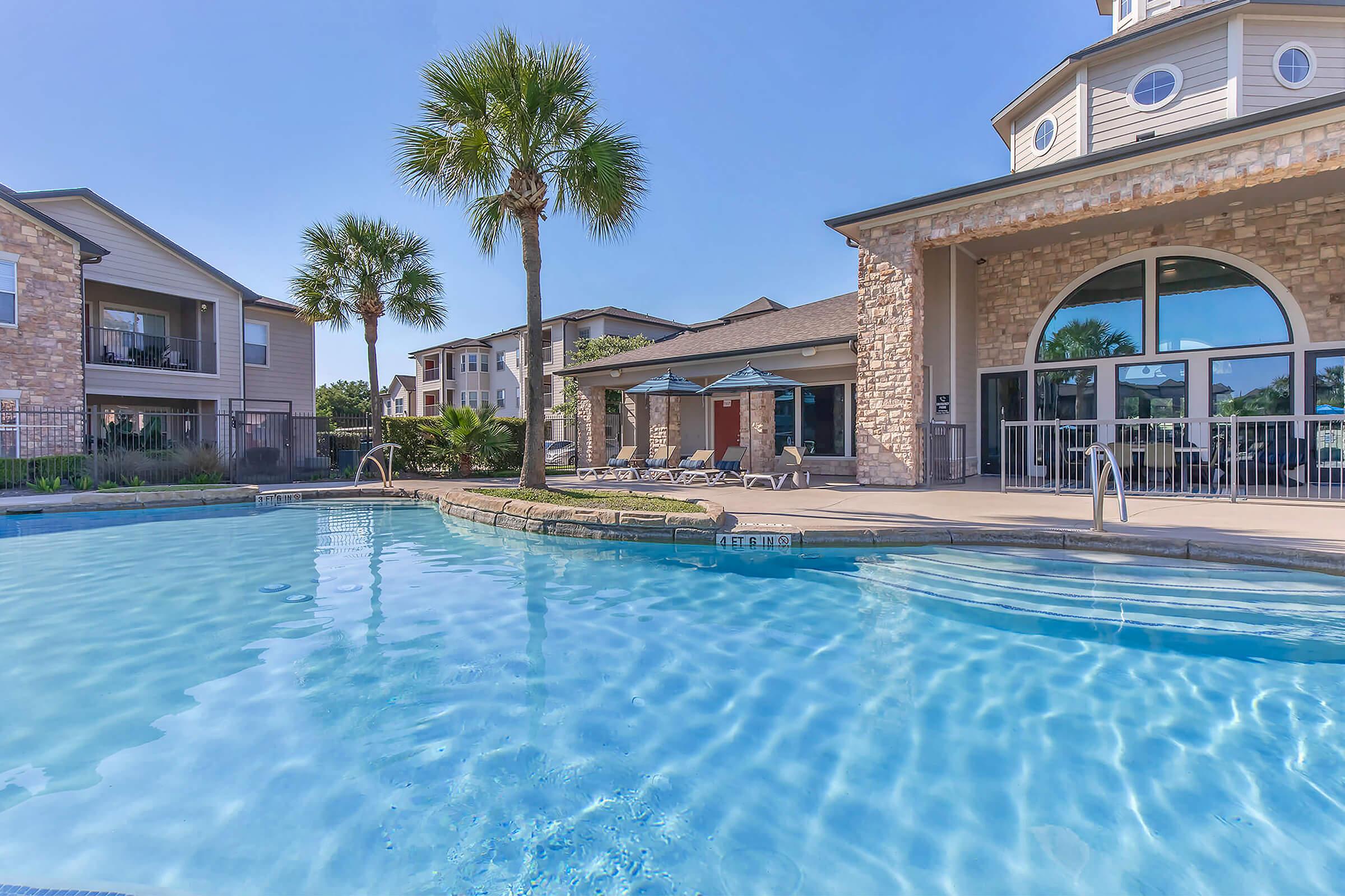 A clear blue swimming pool surrounded by palm trees and lounge chairs, with an apartment building in the background. The setting is sunny and inviting, featuring a patio area with umbrellas and a stone-clad clubhouse. Perfect for relaxation and leisure.