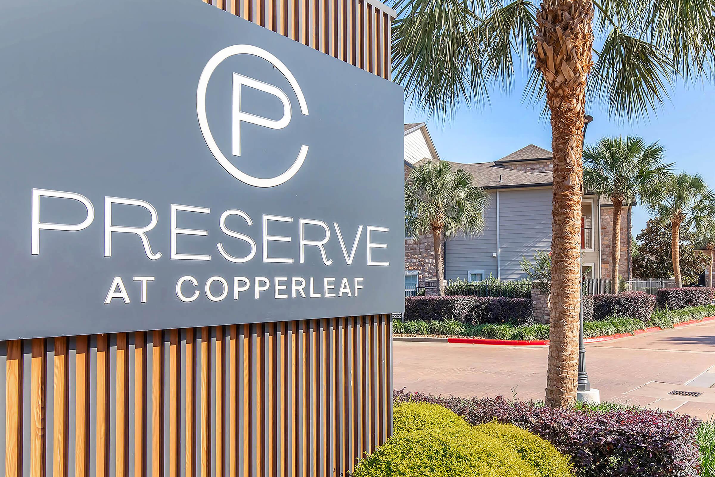 Sign for "Preserve at Copperleaf" featuring a large letter "P" logo. Surrounding landscaping includes palm trees and shrubs, creating a welcoming entrance to an apartment community, with a clear blue sky above.