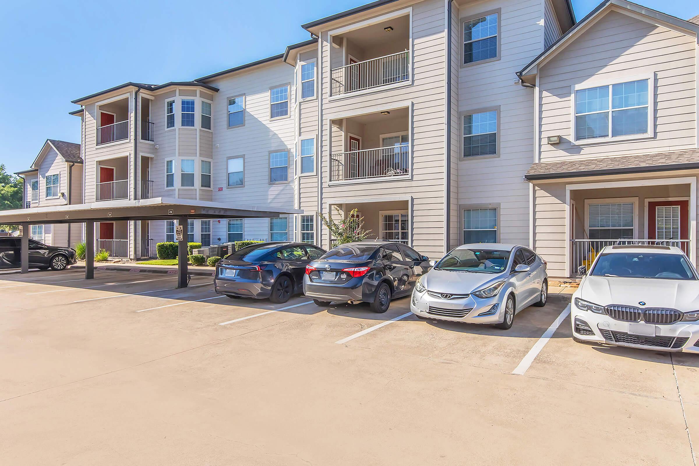 Apartments with a modern design, featuring a three-story building with balconies. Several parked cars are visible in the front, with trees and landscaping enhancing the area. The parking lot is clean and well-maintained, indicating a welcoming residential environment.
