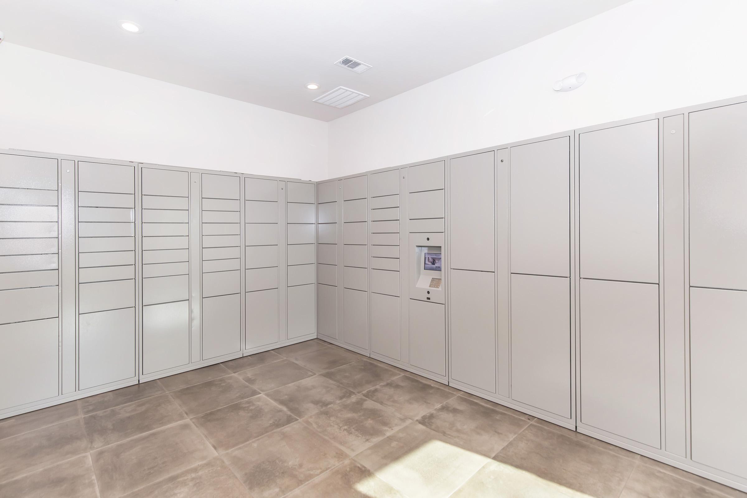 Empty package delivery room featuring multiple grey lockers against white walls, with a floor of large gray tiles. A small kiosk or screen is present in the middle, likely for retrieving packages. The room is well-lit with overhead lights.