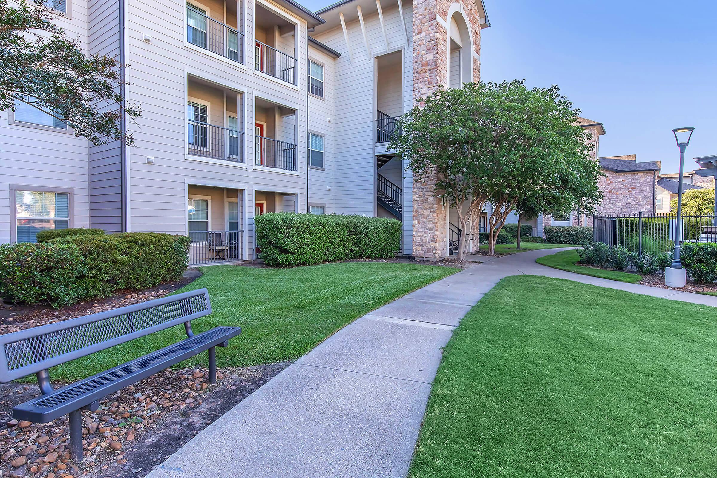 A well-maintained apartment complex featuring multiple stories, with a walkway leading past manicured lawns and shrubs. A black bench sits along the path, and there are trees providing shade. The building has a stone and beige exterior, with balconies visible on the upper floors.