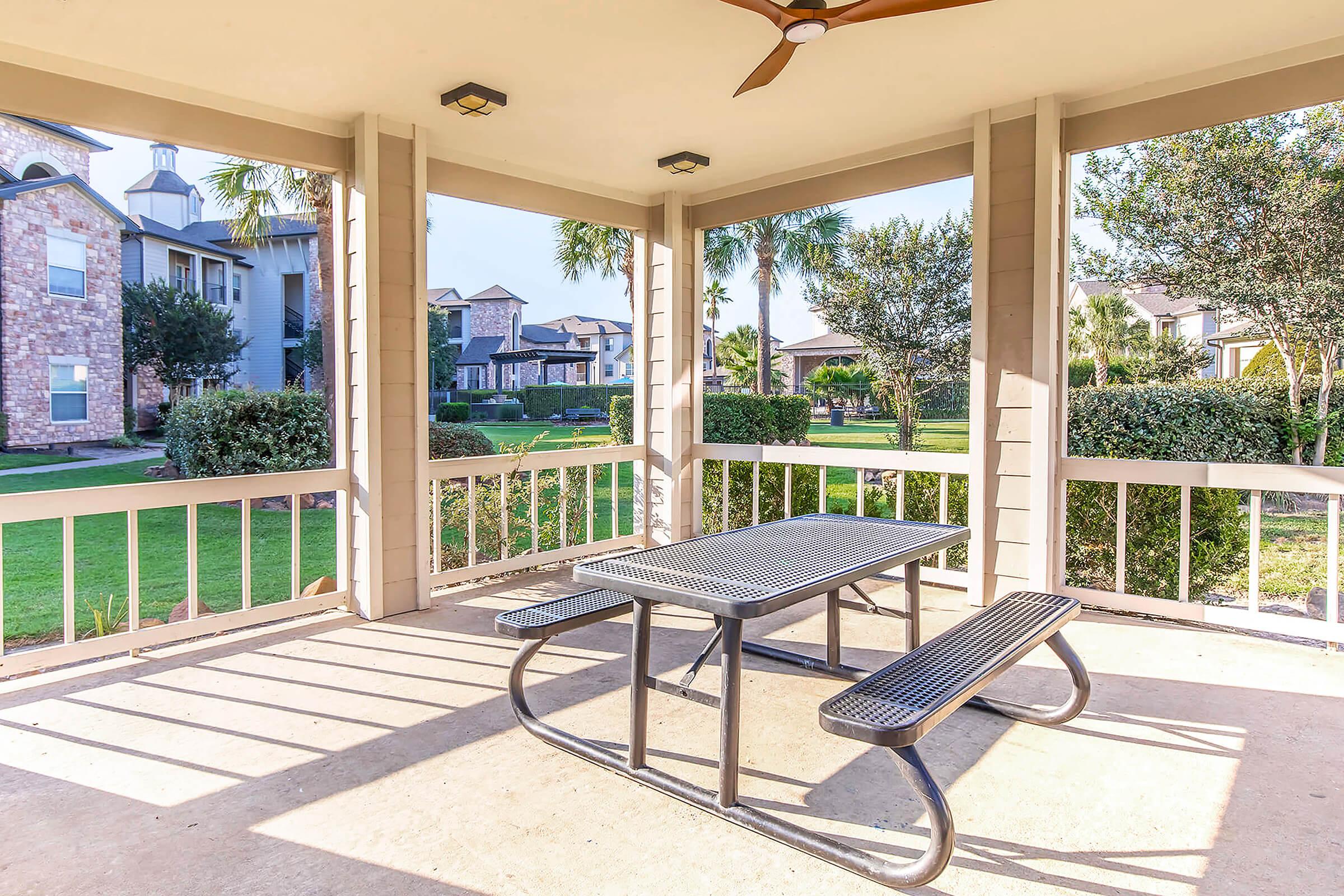 A shaded outdoor gazebo with a picnic table and benches, surrounded by lush green lawns and landscaping. Nearby buildings are visible in the background, and palm trees add a tropical feel to the setting. Sunlight filters through, creating a bright and inviting atmosphere.