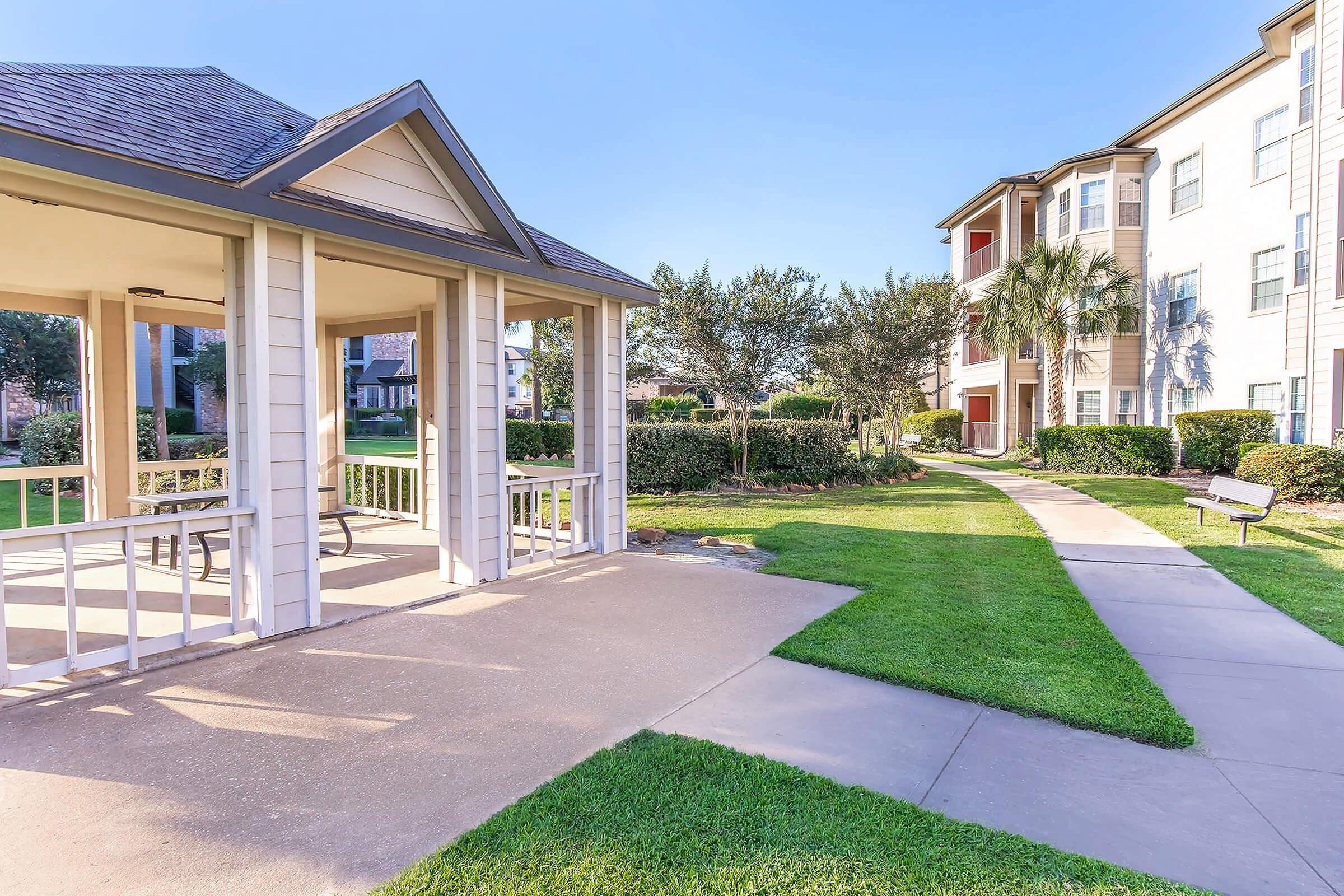 A landscaped area featuring a gazebo with benches, surrounded by lush green grass and shrubs. A clear pathway leads gently through the space, with a multi-story apartment building visible in the background and palm trees adding to the aesthetic appeal. The sky is bright and clear.