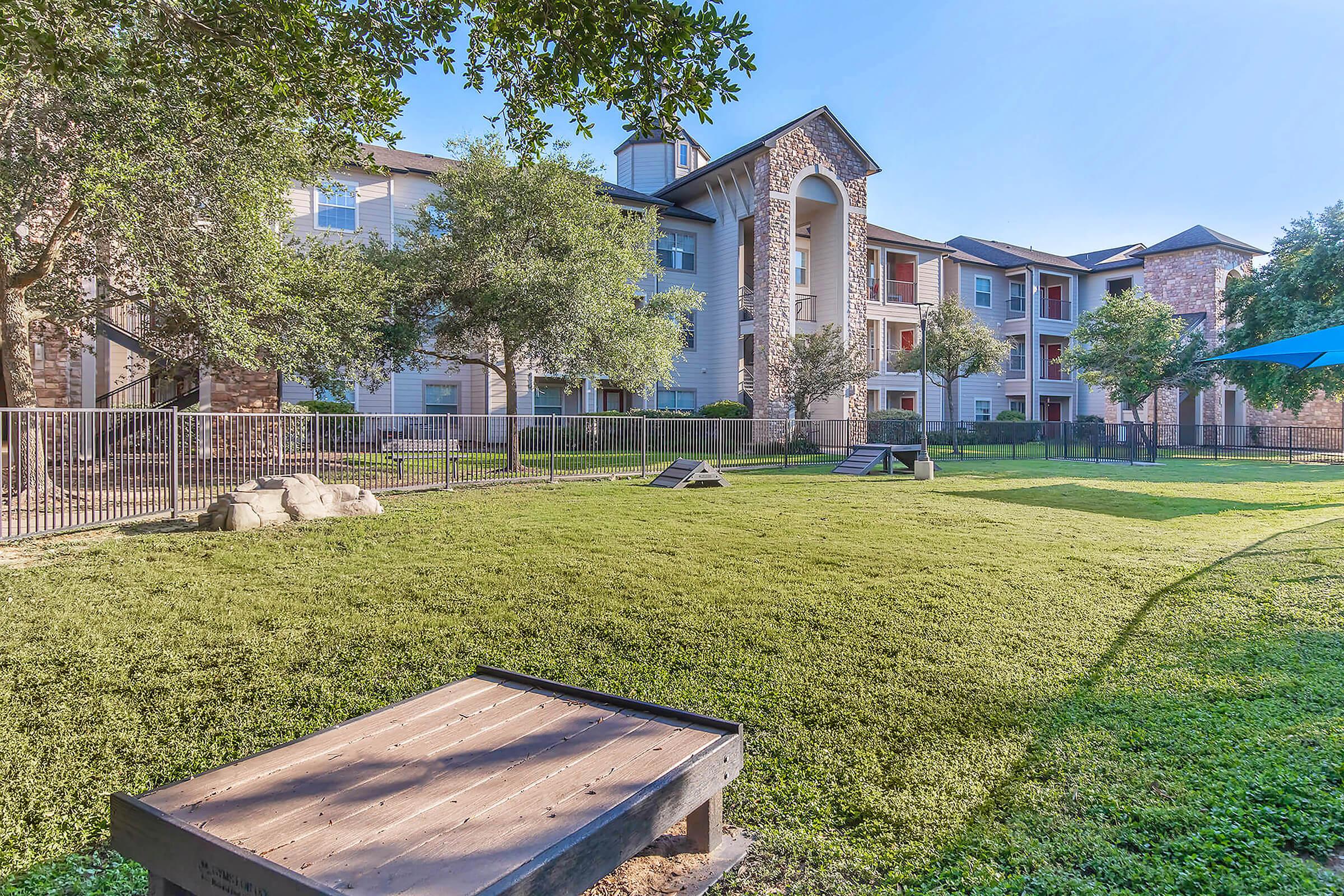 A sunny outdoor area featuring a well-maintained lawn with a small dog park, including two agility structures. In the background, a multi-story residential building is visible, surrounded by trees and a fence. The scene conveys a welcoming, pet-friendly environment.
