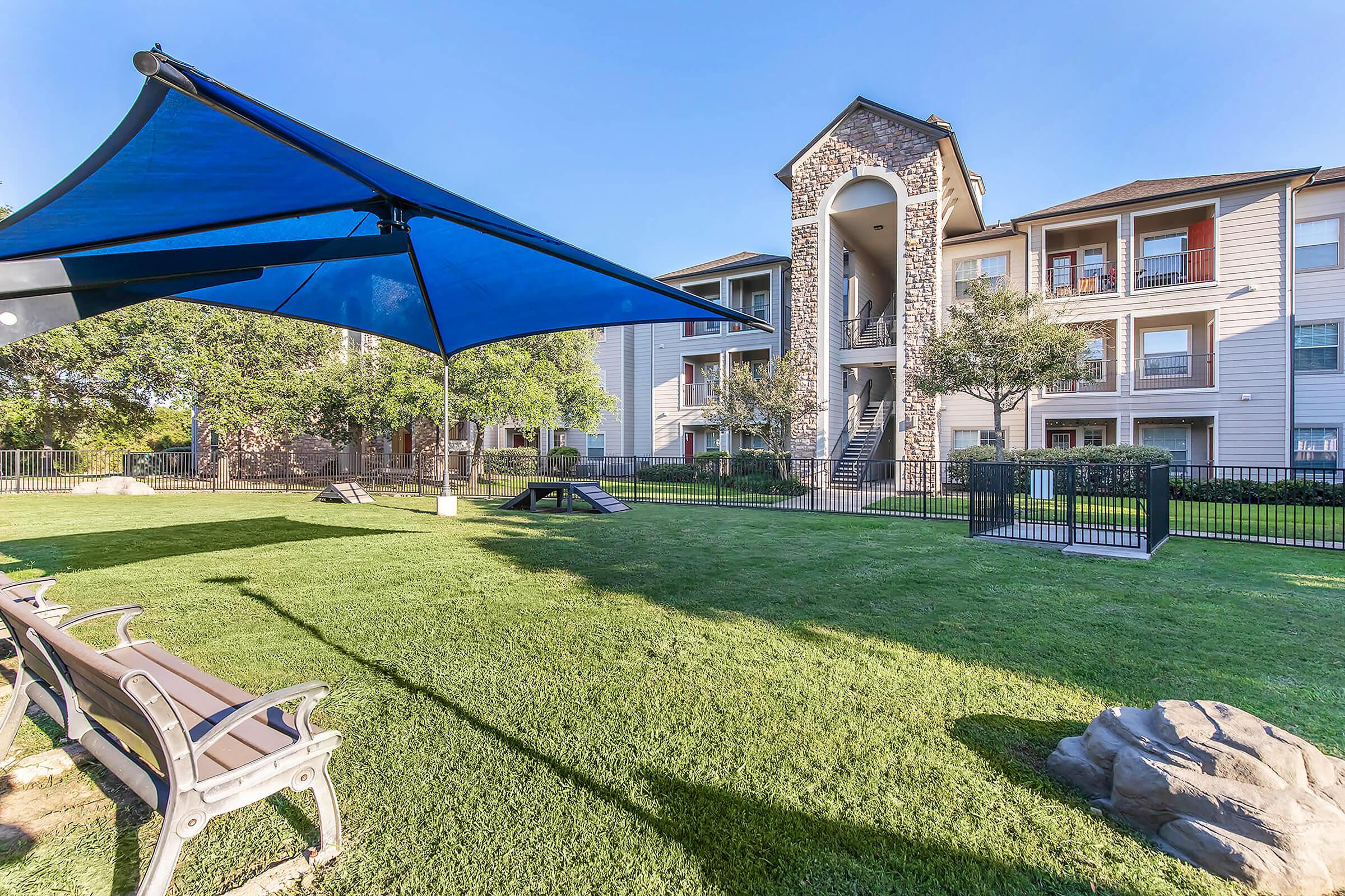 View of a well-maintained apartment complex featuring a grassy courtyard, benches for seating, and a large blue shade structure. The buildings are modern with multiple stories and balconies. Lush greenery surrounds the area, providing a pleasant outdoor space for residents.