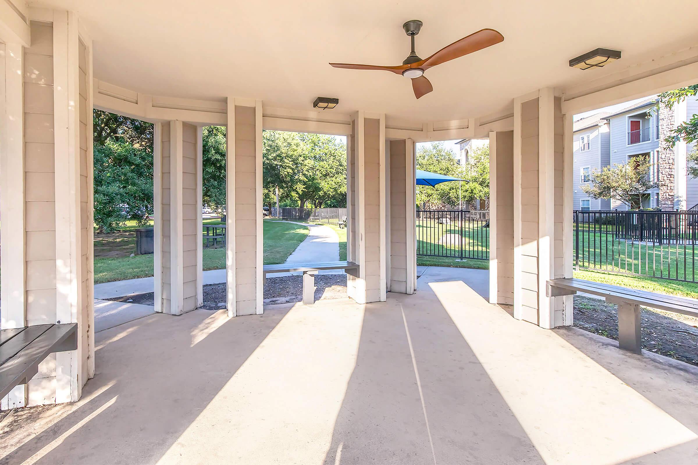 A sheltered gazebo with a fan, featuring open sides and visible beams. Sunlight casts shadows on the concrete floor, while trees and a walking path are seen in the background, creating a serene outdoor setting. Benches are located on either side of the structure.