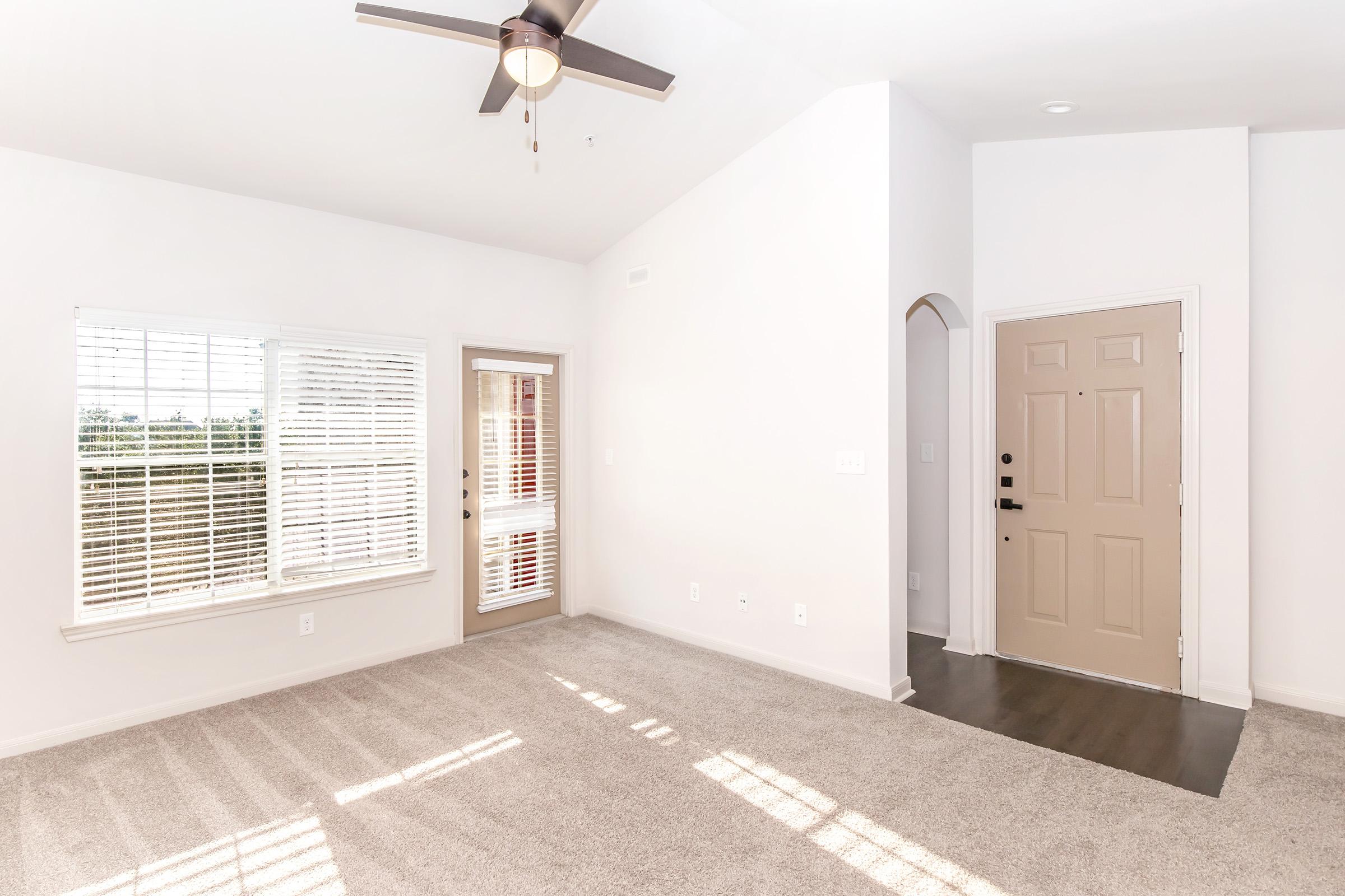 A bright, spacious living area with light-colored walls and plush carpeting. A window with white blinds lets in natural light, and there's a ceiling fan for air circulation. A door leads to the outside, while another door opens to a hallway. The overall ambiance is inviting and modern.