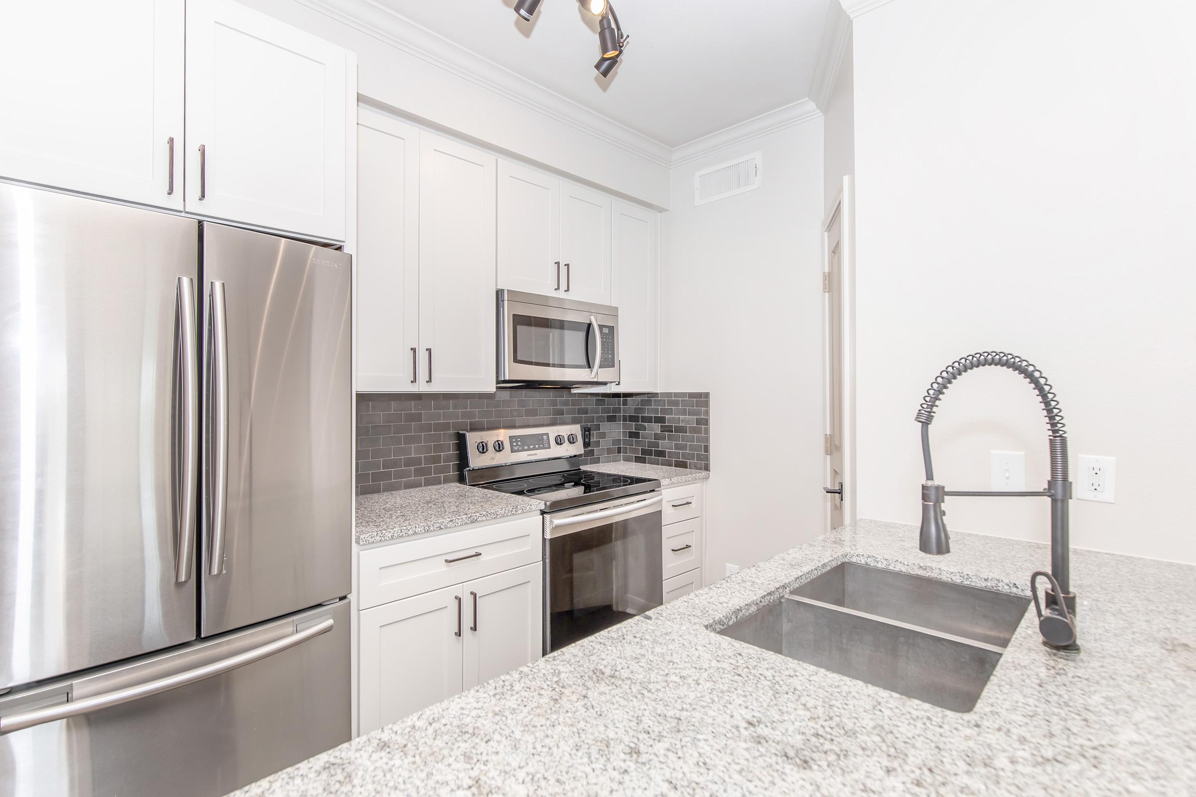 Modern kitchen featuring stainless steel appliances, including a refrigerator, microwave, and oven. The space has light-colored cabinetry, a granite countertop, and a stainless steel sink with a stylish faucet. The backsplash consists of dark tiles, creating a contrast with the cabinetry.