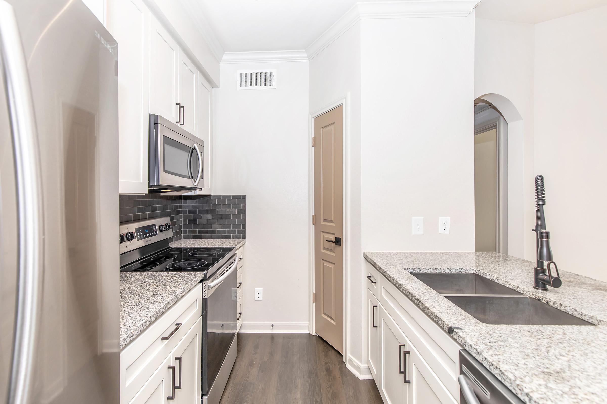 A modern kitchen featuring stainless steel appliances, including an oven and microwave, with a large granite countertop. The design includes white cabinets, a sleek kitchen sink, and a subtle gray backsplash. A doorway leads to another room, highlighting an open and spacious layout.