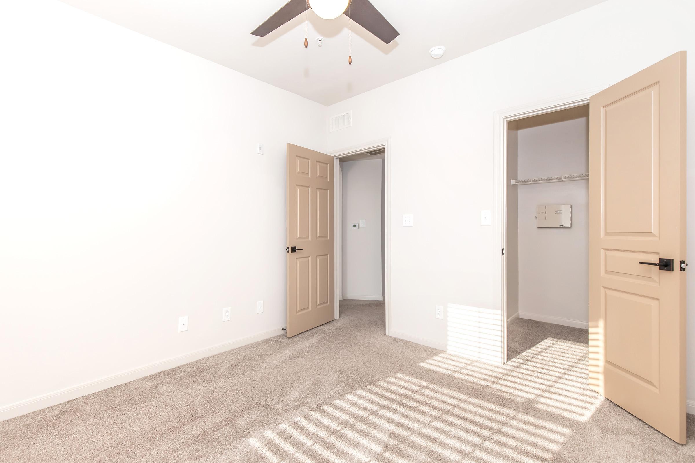 Empty room with light-colored walls and carpeted flooring. A ceiling fan is installed, and two doors are open: one leading to a closet and the other to an adjoining room. Sunlight casts shadows on the floor from the window, creating a bright, inviting atmosphere.