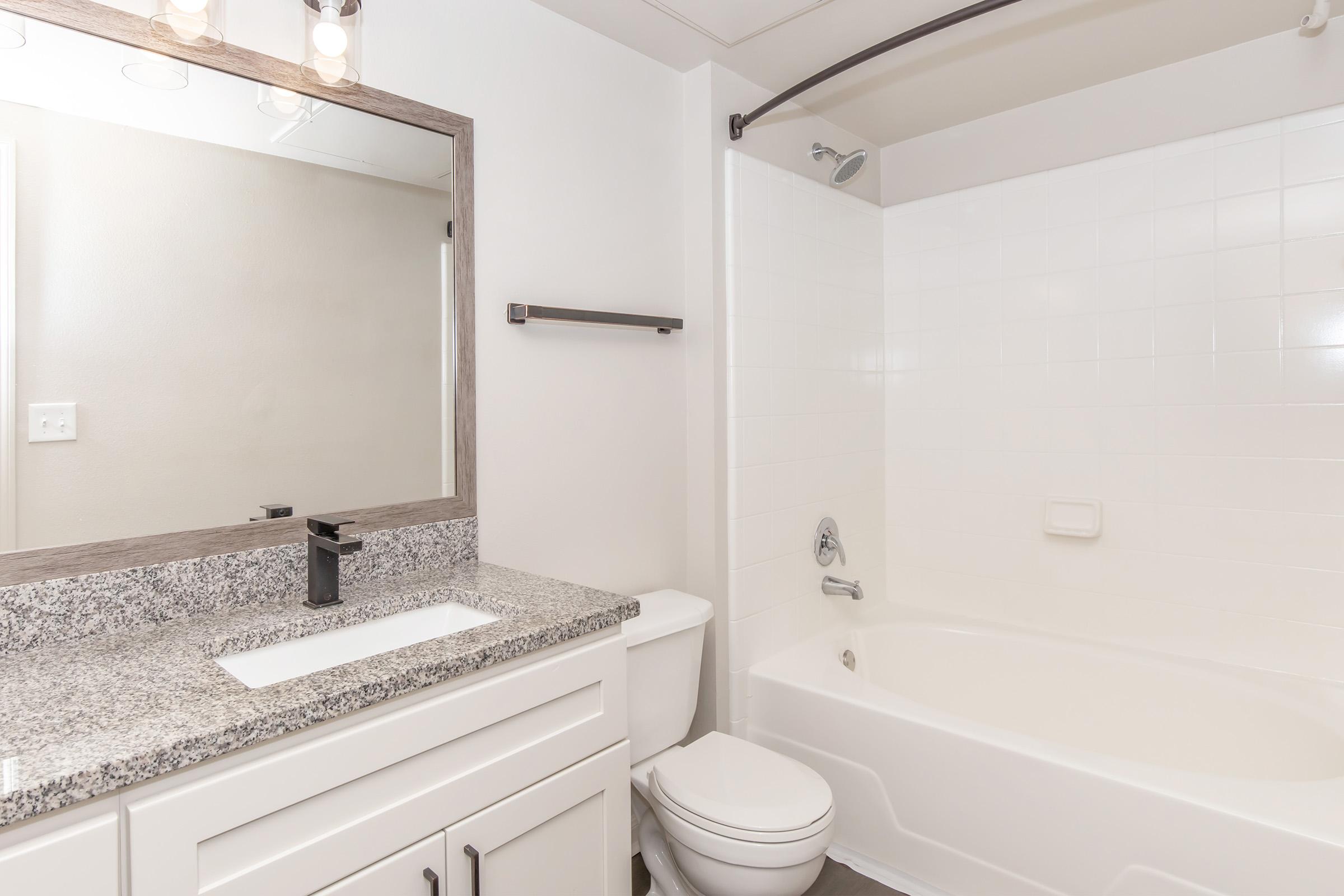 Modern bathroom featuring a white tiled bathtub with a shower, a white vanity with a gray granite countertop, a sleek black faucet, and a large mirror above the sink. The walls are light-colored, creating a bright and clean aesthetic. A towel bar is installed next to the bathtub.