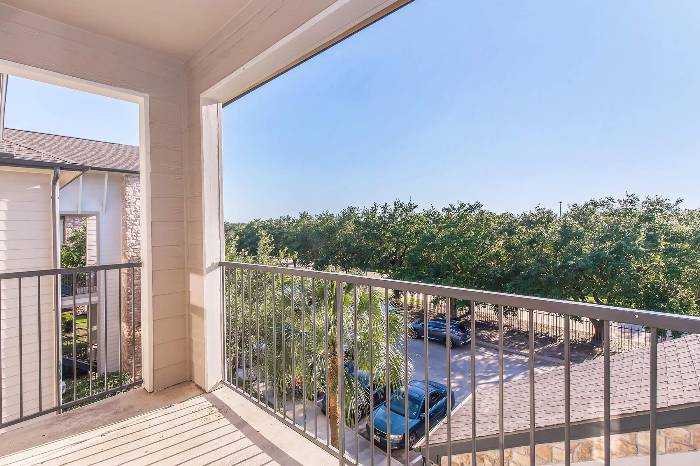 A balcony view overlooking lush greenery and parked cars. The scene features a clear blue sky and a spacious outdoor area, creating a serene atmosphere. The balcony has a metal railing and wooden flooring, providing a comfortable space to enjoy the surroundings.