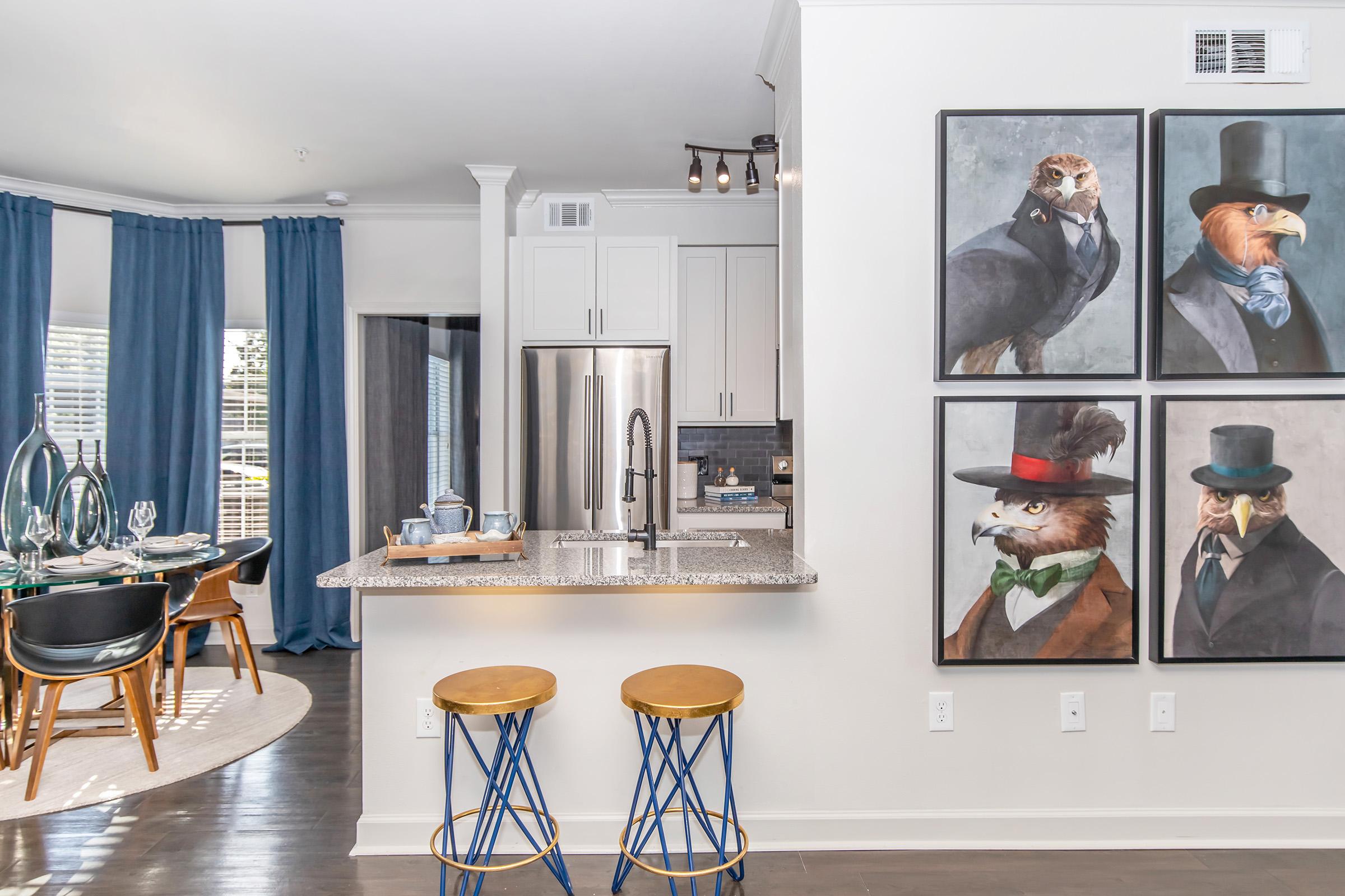 Modern kitchen and dining area with a light color scheme. The kitchen features a granite countertop and stainless steel appliances. Blue curtains frame the window, and four unique portraits of anthropomorphic birds in suits adorn the wall. Two stylish wooden stools are positioned at the kitchen counter.