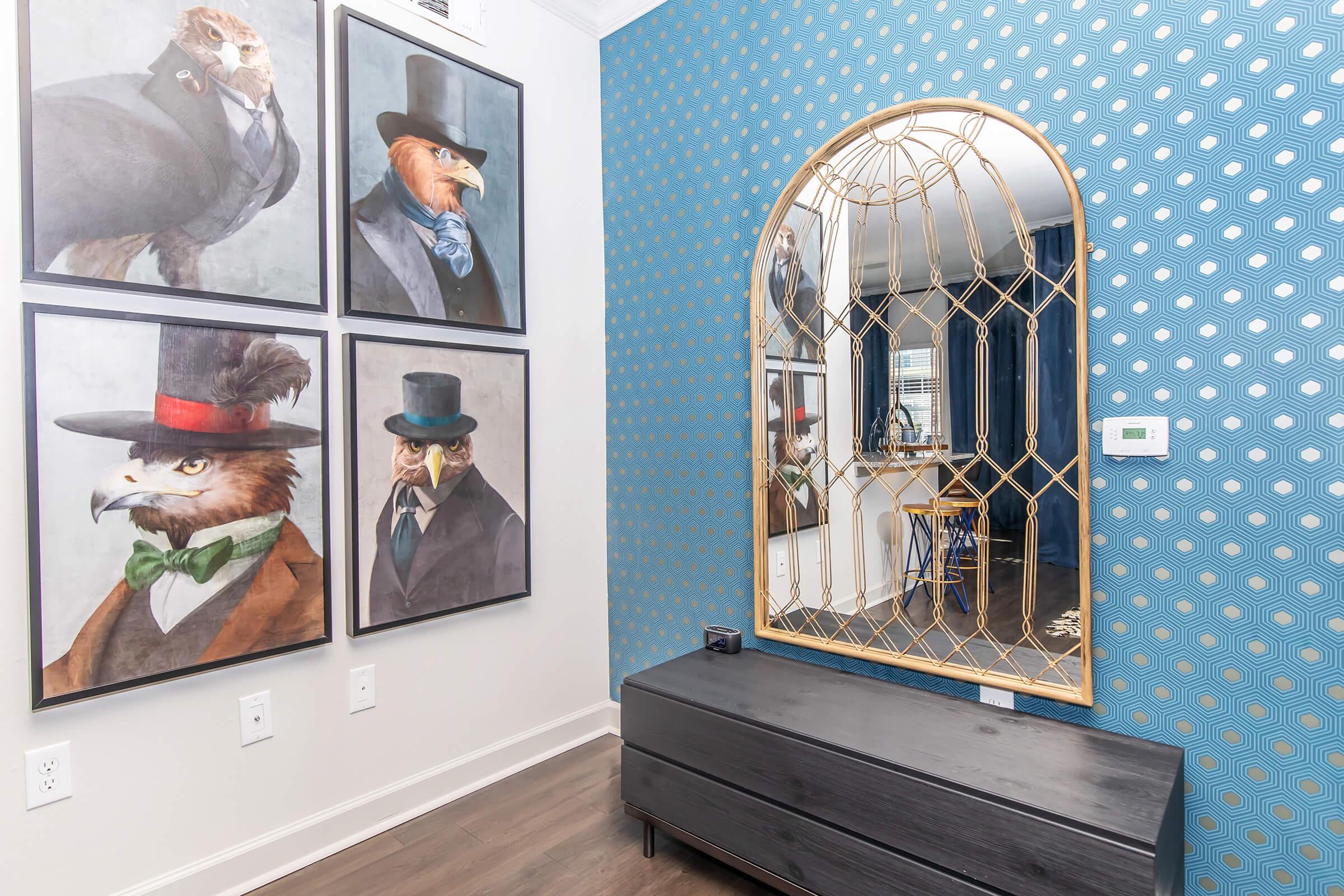 A stylish interior featuring four framed animal portraits in formal attire, adorned with top hats and bow ties, hung on a blue wall with a circular patterned design. In front, there is a sleek black bench and a large, ornate mirror reflecting the space. A glimpse of a kitchen can be seen in the background.