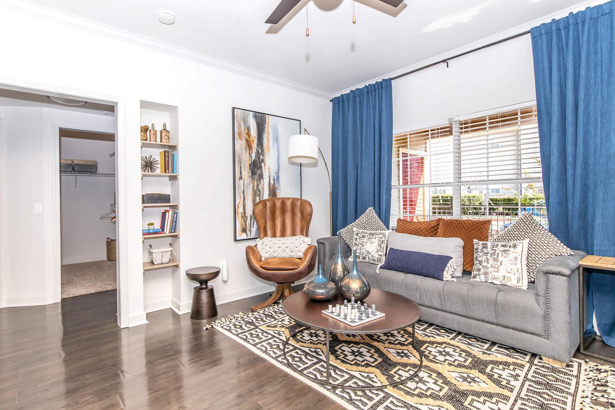 A cozy living room featuring a gray sofa with decorative pillows, a brown armchair, and a round coffee table with a chess set. The space is adorned with blue curtains, a large artwork on the wall, and a bookshelf. A closet door is visible in the background, and the flooring is dark wood.