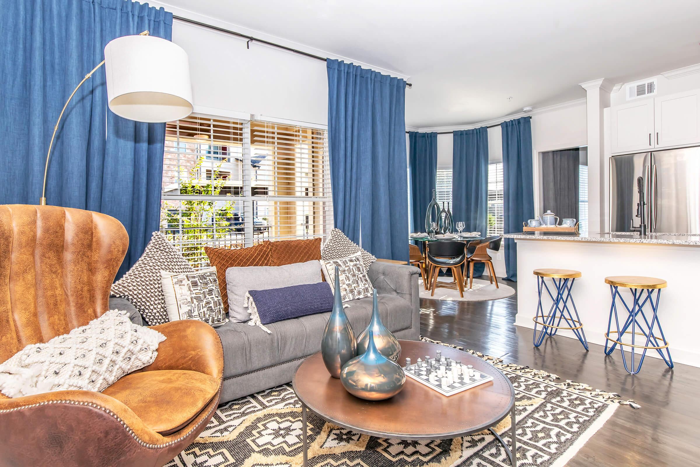 A cozy living room featuring a brown leather armchair, a gray sofa adorned with various textured pillows, and a round coffee table with decorative items. The space has large windows draped with blue curtains, and a dining area visible in the background, highlighted by modern seating and a stylish kitchen.