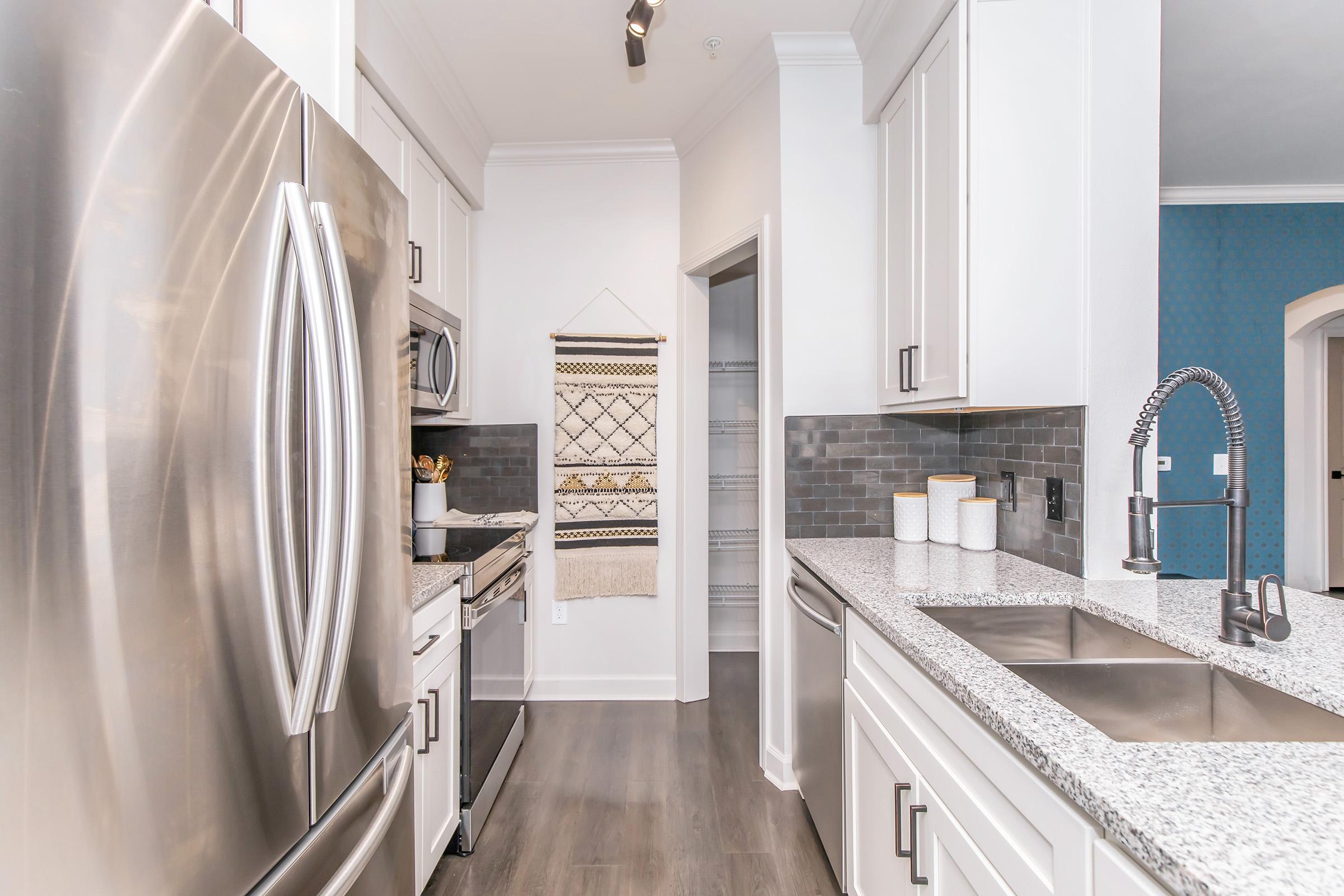 A modern kitchen featuring stainless steel appliances, including a refrigerator and oven. The countertops are gray speckled granite, with a double sink and a sleek faucet. Light-colored cabinets line the kitchen, and a pantry door is visible in the background. The walls showcase a light, neutral color.