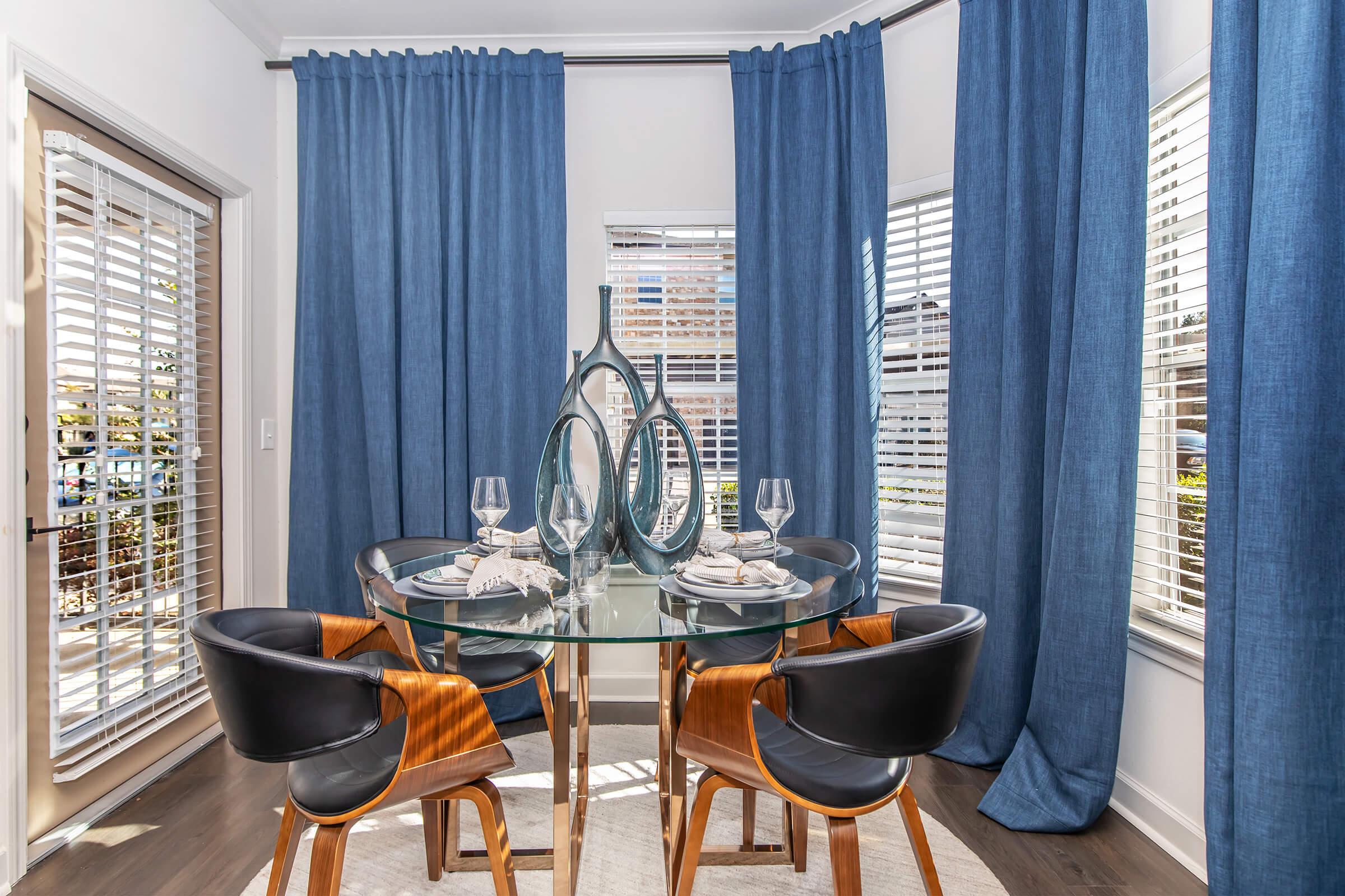 A modern dining area featuring a round glass table with elegant table settings, including plates and wine glasses. Surrounded by stylish wooden chairs, the space is adorned with blue curtains and has abundant natural light coming through the window.