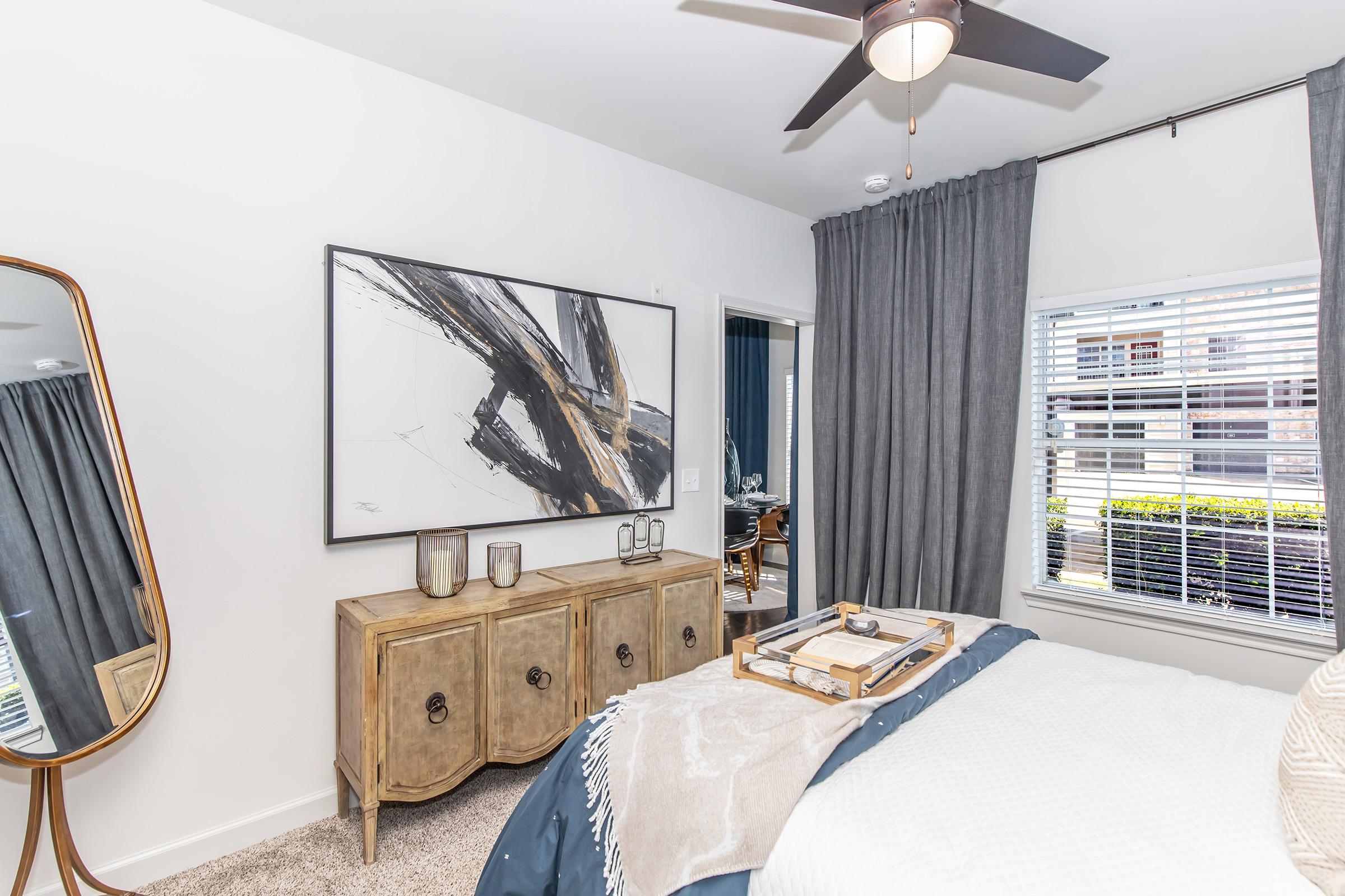 A modern bedroom featuring a large mirror, a stylish dresser with decorative items, and a painting with abstract art on the wall. The room has gray curtains, a ceiling fan, and a cozy bed with decorative pillows, creating a warm and inviting atmosphere.
