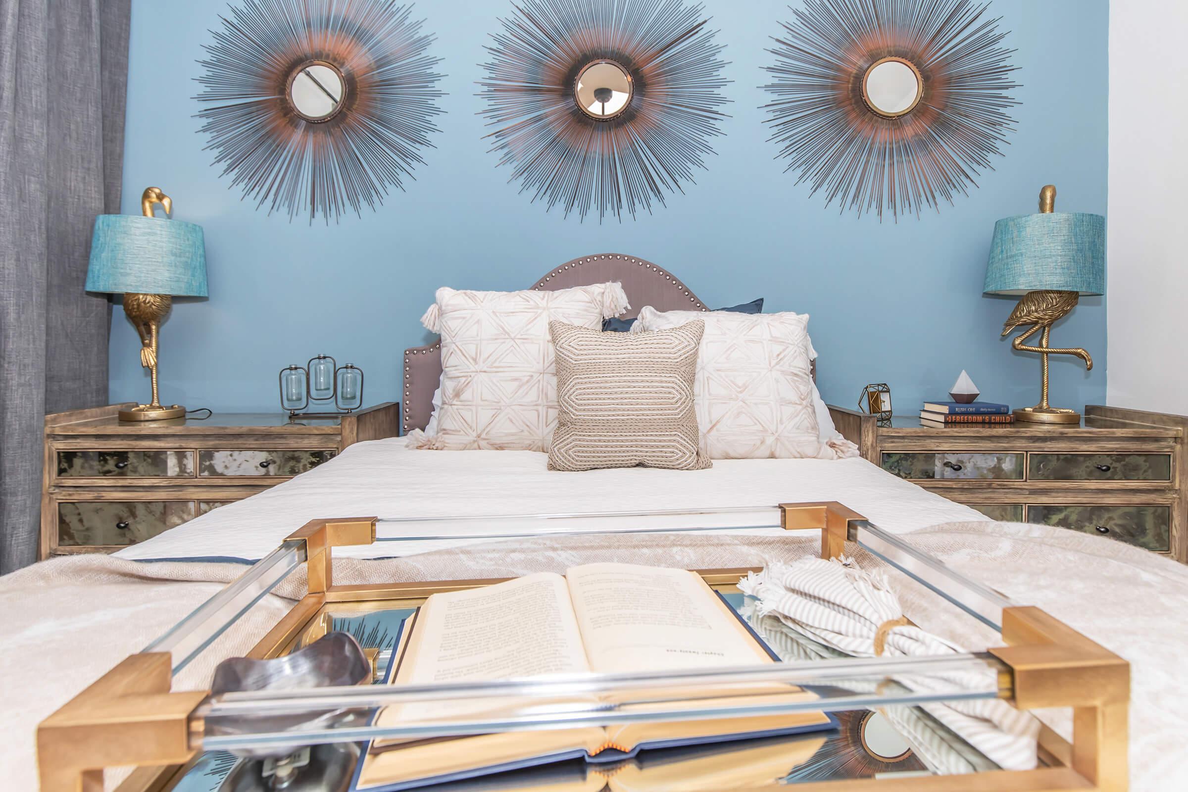 A stylish bedroom featuring a large bed with decorative pillows and a textured throw. The background is painted light blue, accented by three sunburst mirrors. There are bedside tables with teal lamps, and a tray on the bed holds a book and decorative items, contributing to a cozy and elegant atmosphere.
