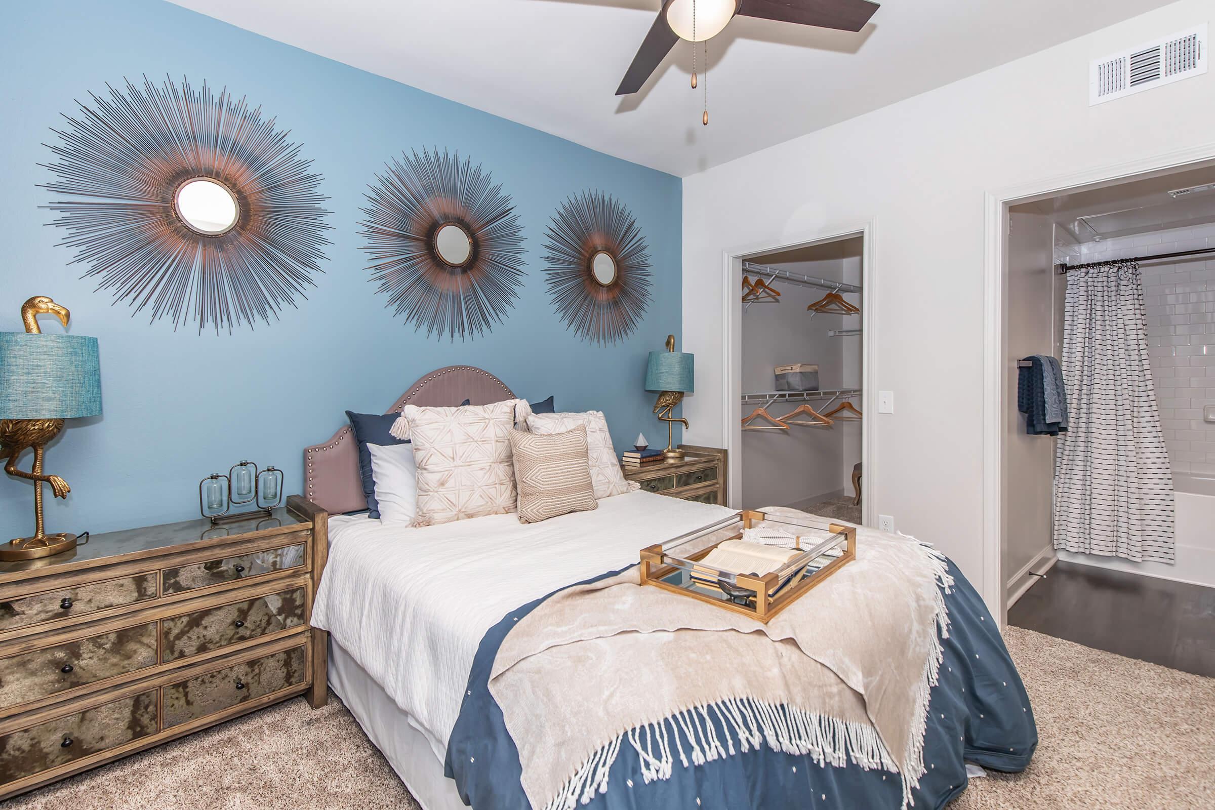 A stylish bedroom featuring a light blue accent wall adorned with three sunburst mirrors. The bed is neatly made with a beige and white throw blanket and decorative pillows. Beside the bed, there are two stylish lamps. A dresser with a mirror is against one wall, and a closet with hanging space is visible in the background.