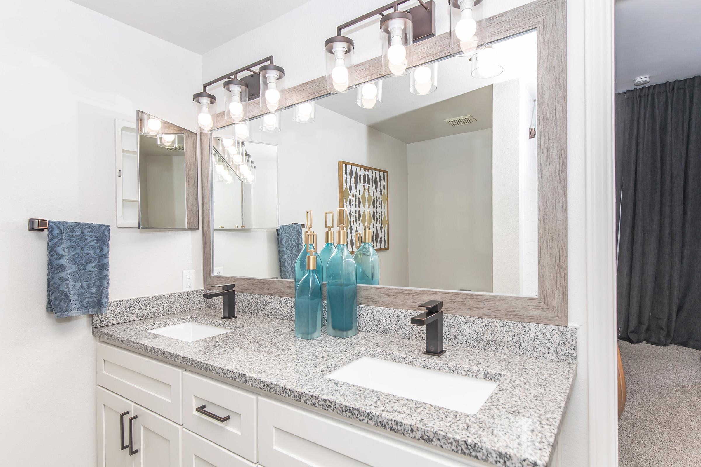 A modern bathroom featuring a dual sink vanity with a granite countertop, large mirror, and decorative blue glass vases. The lighting consists of several exposed bulbs above the mirror. A blue towel hangs on the left, and there is a dark curtain in the background, adding a contemporary touch to the space.