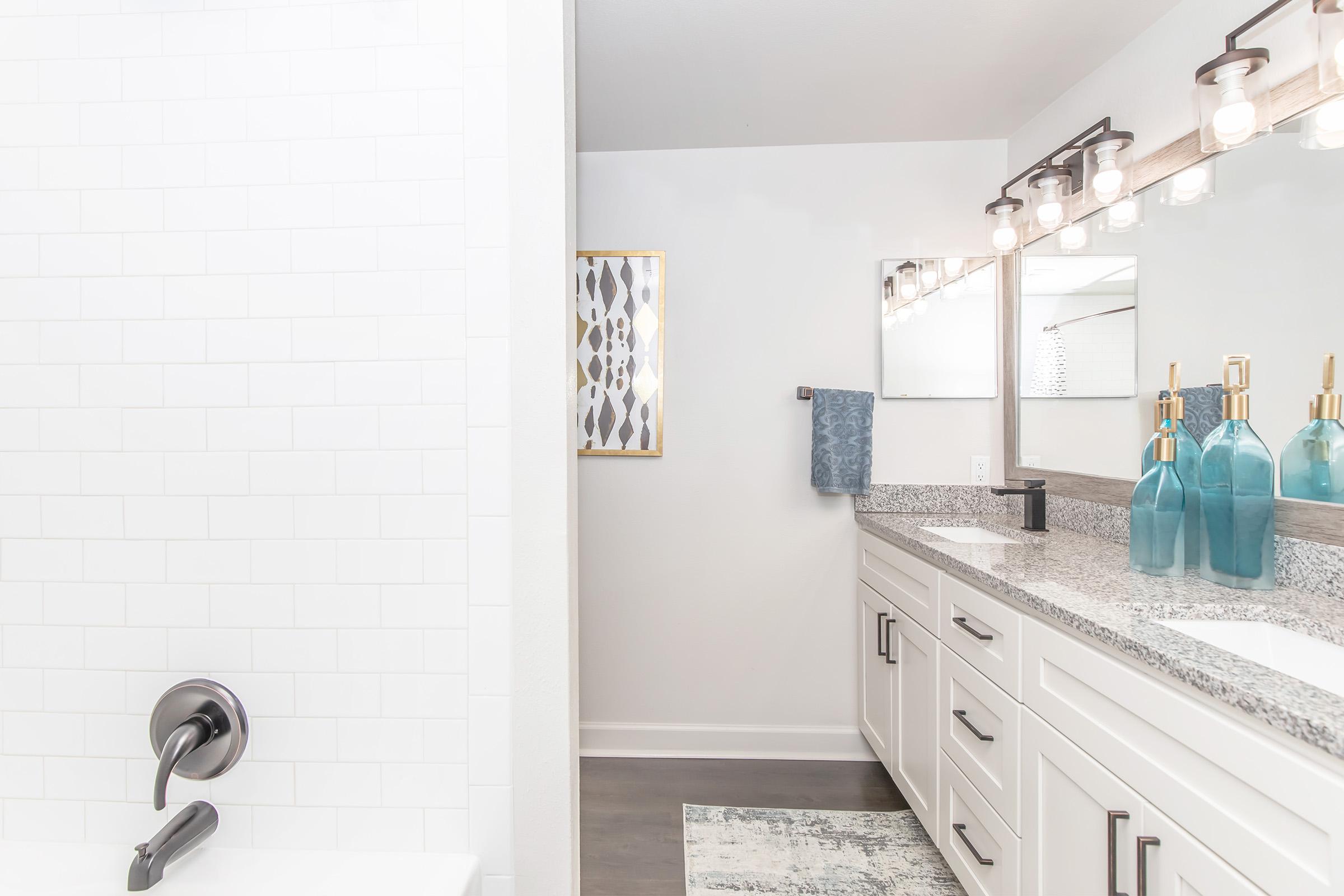 Modern bathroom with a white tiled shower and a double vanity. The vanity features a gray countertop, white cabinetry, and a large mirror with stylish lighting above. Decorative bottles in shades of blue sit on the counter, and a patterned artwork is displayed on the wall. A towel hangs beside the mirror.