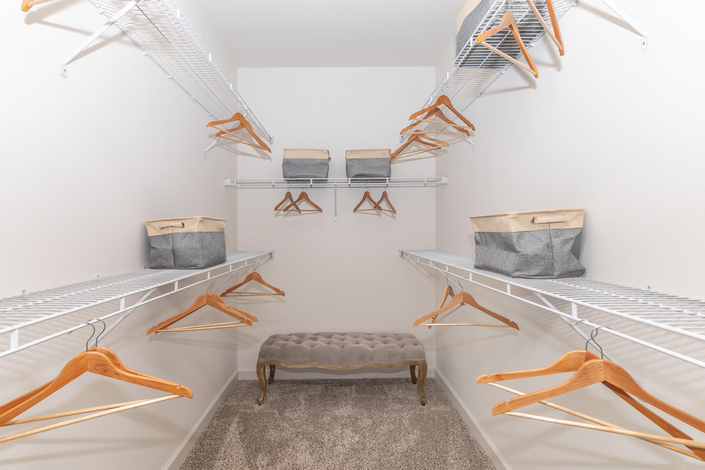 A spacious walk-in closet featuring white wire shelves lined with wooden hangers. Two fabric storage bins are placed on the upper shelves, and a plush, upholstered bench sits in the center on a soft carpeted floor. The walls are plain and unadorned, creating a clean, organized look.