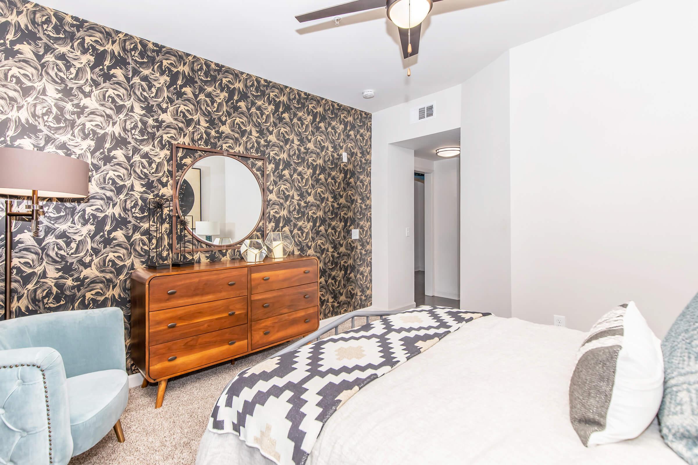 A stylish bedroom featuring a patterned accent wall, a round mirror above a wooden dresser, and a plush bed with a decorative blanket. A cozy armchair is in the corner, and soft lighting creates a warm ambiance. The floor is carpeted, adding to the comfort of the space.