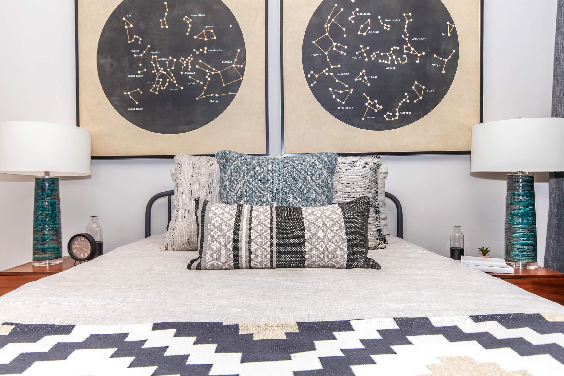 A cozy bedroom featuring a bed with various decorative pillows and a patterned blanket. Behind the bed are two large framed art pieces depicting constellations. On either side of the bed, there are stylish lamps with blue bases.