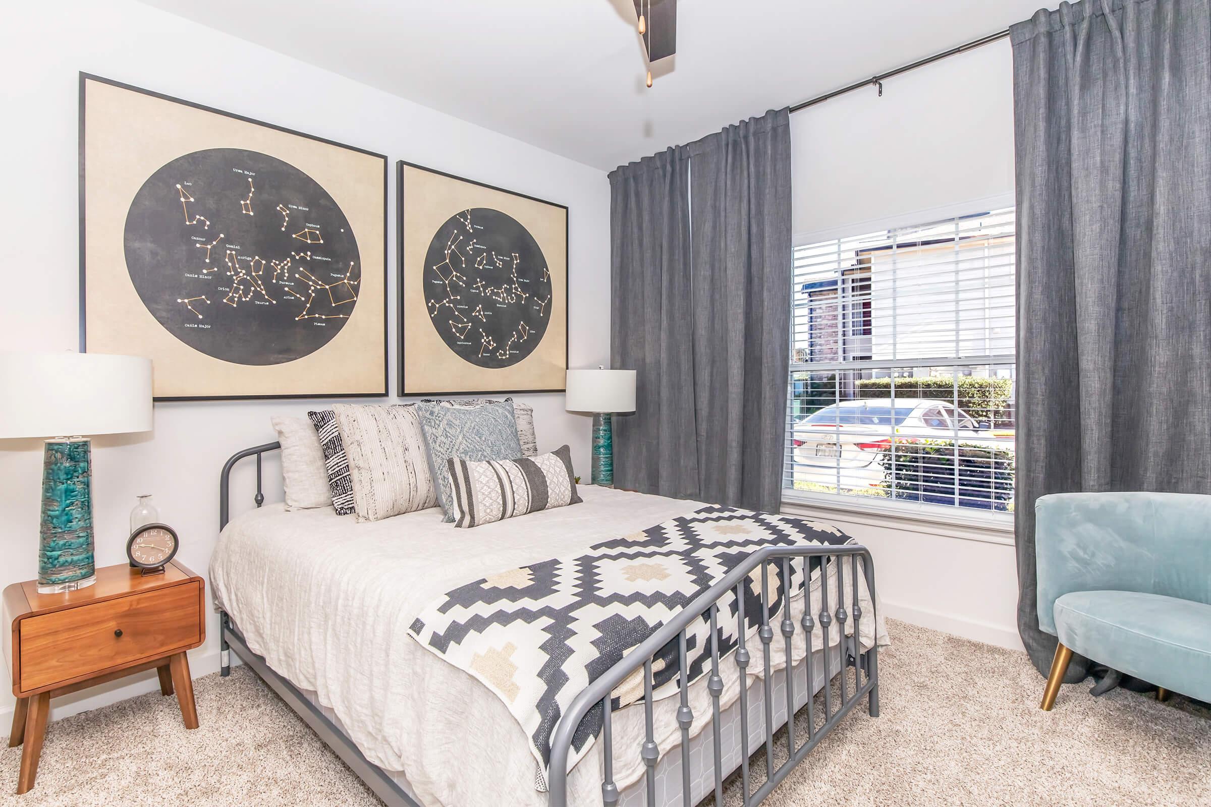 A cozy bedroom featuring a metal bed frame with a light-colored quilt and decorative pillows. Two large framed star maps adorn the wall above the bed. There are two bedside lamps, a wooden nightstand, and a teal accent chair. Sheer gray curtains frame a window that lets in natural light.
