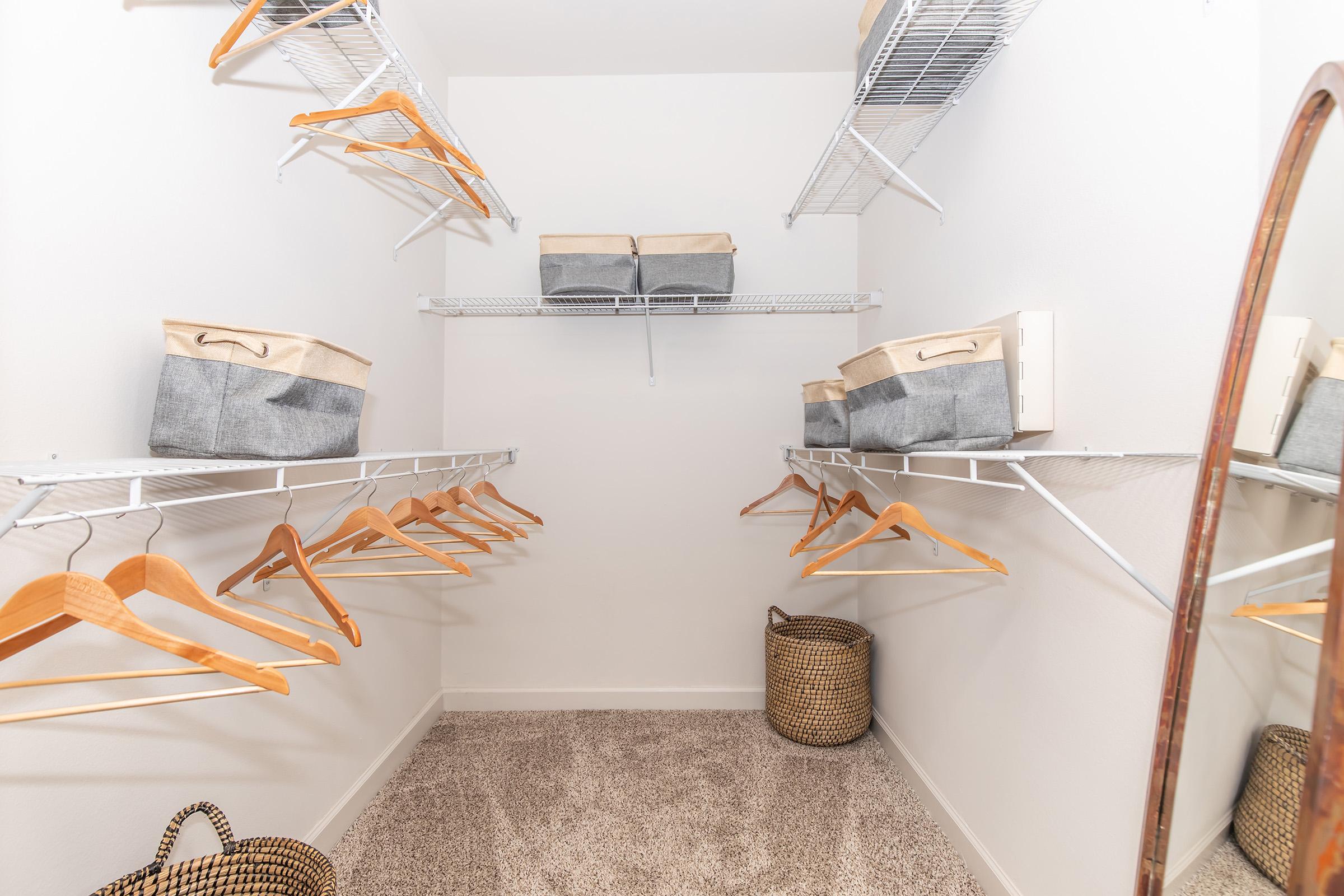 A walk-in closet with white walls and carpeted flooring. It features wire shelving with several wooden hangers and storage bins. A woven basket is placed on the floor, and a full-length mirror is visible on the right side of the image. The space is organized and well-lit.