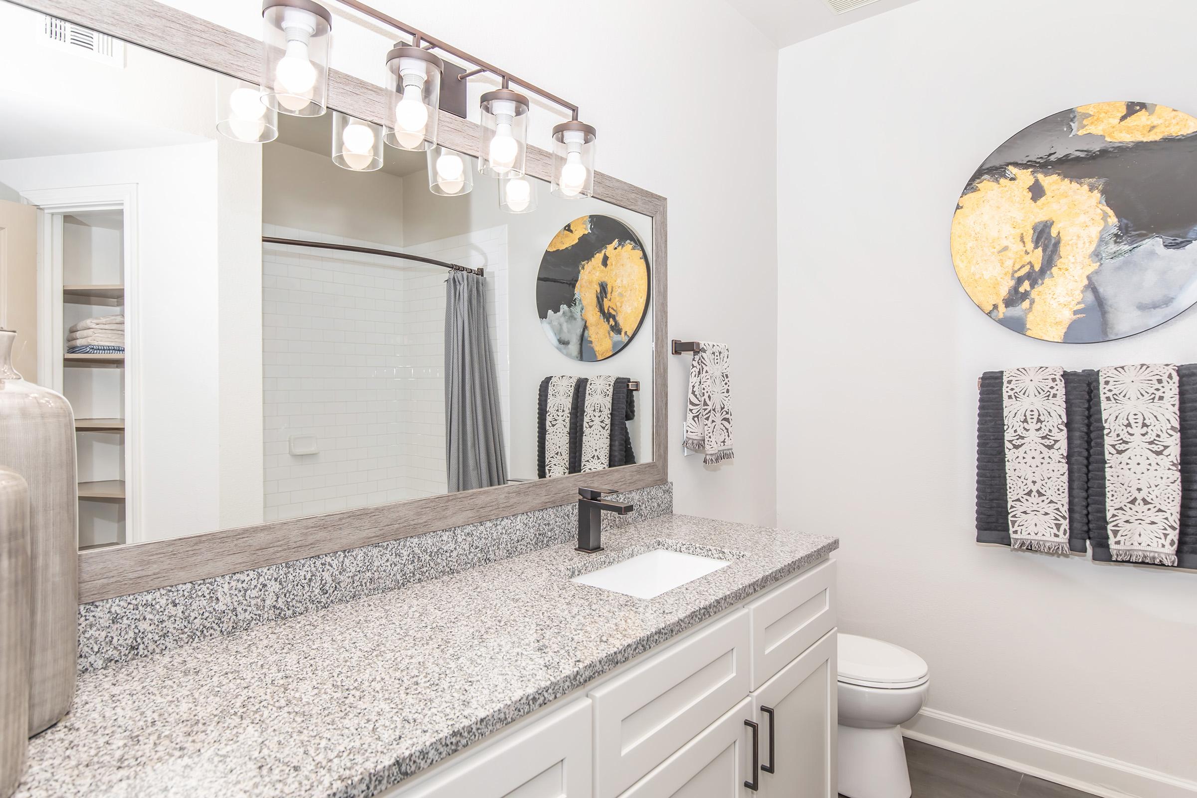 A modern bathroom featuring a large mirror with a stylish light fixture above. The countertop is made of granite, and there are neatly organized towels hanging on a rack. A round decorative wall piece resembling a map is mounted above the sink. The shower area is partially visible on the left.