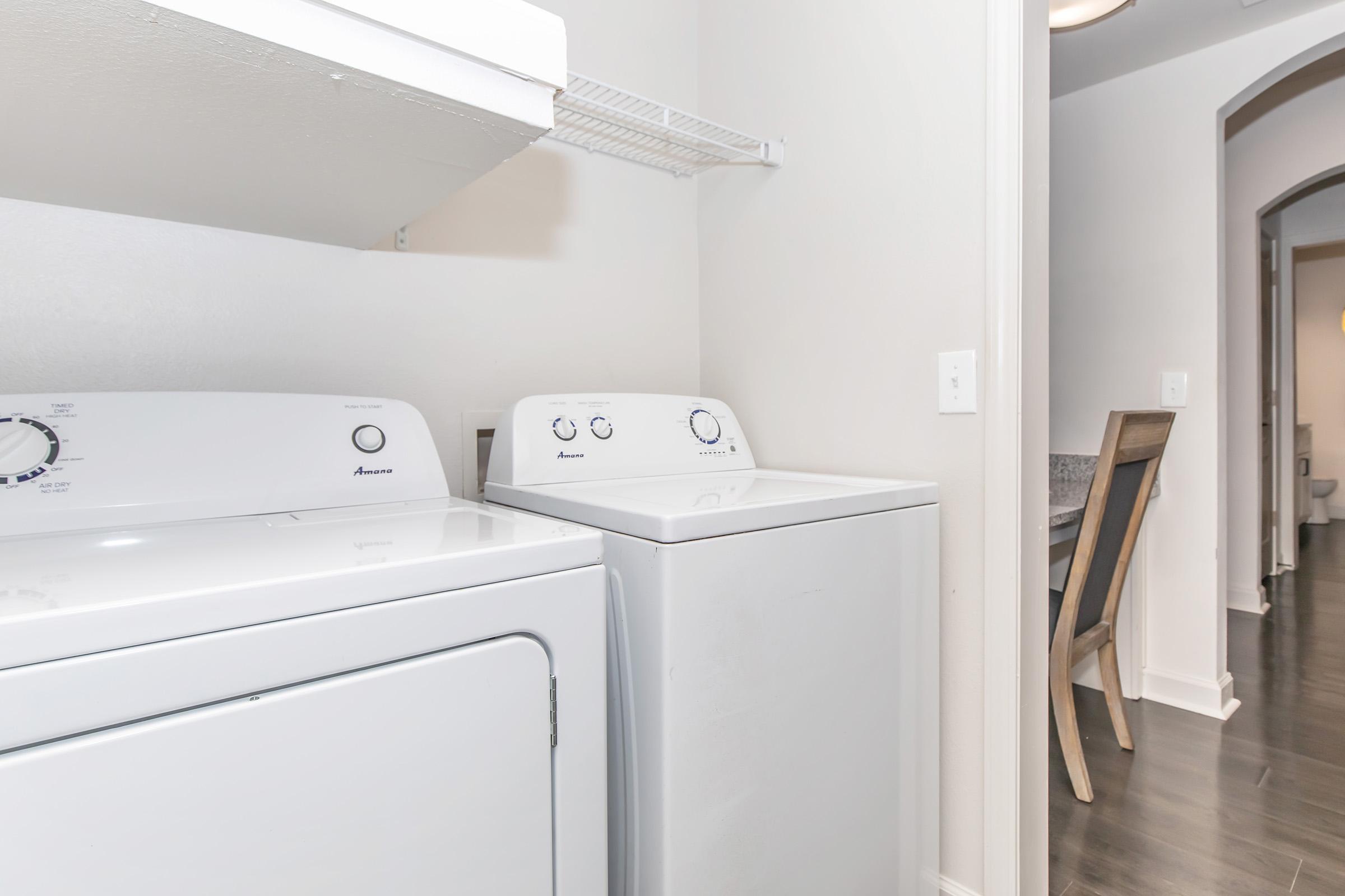 A clean and organized laundry room featuring a white washing machine and dryer side by side. The machines are placed against a light-colored wall with a shelf above for storage. In the background, there is a glimpse of a hallway leading to a dining area with a chair.